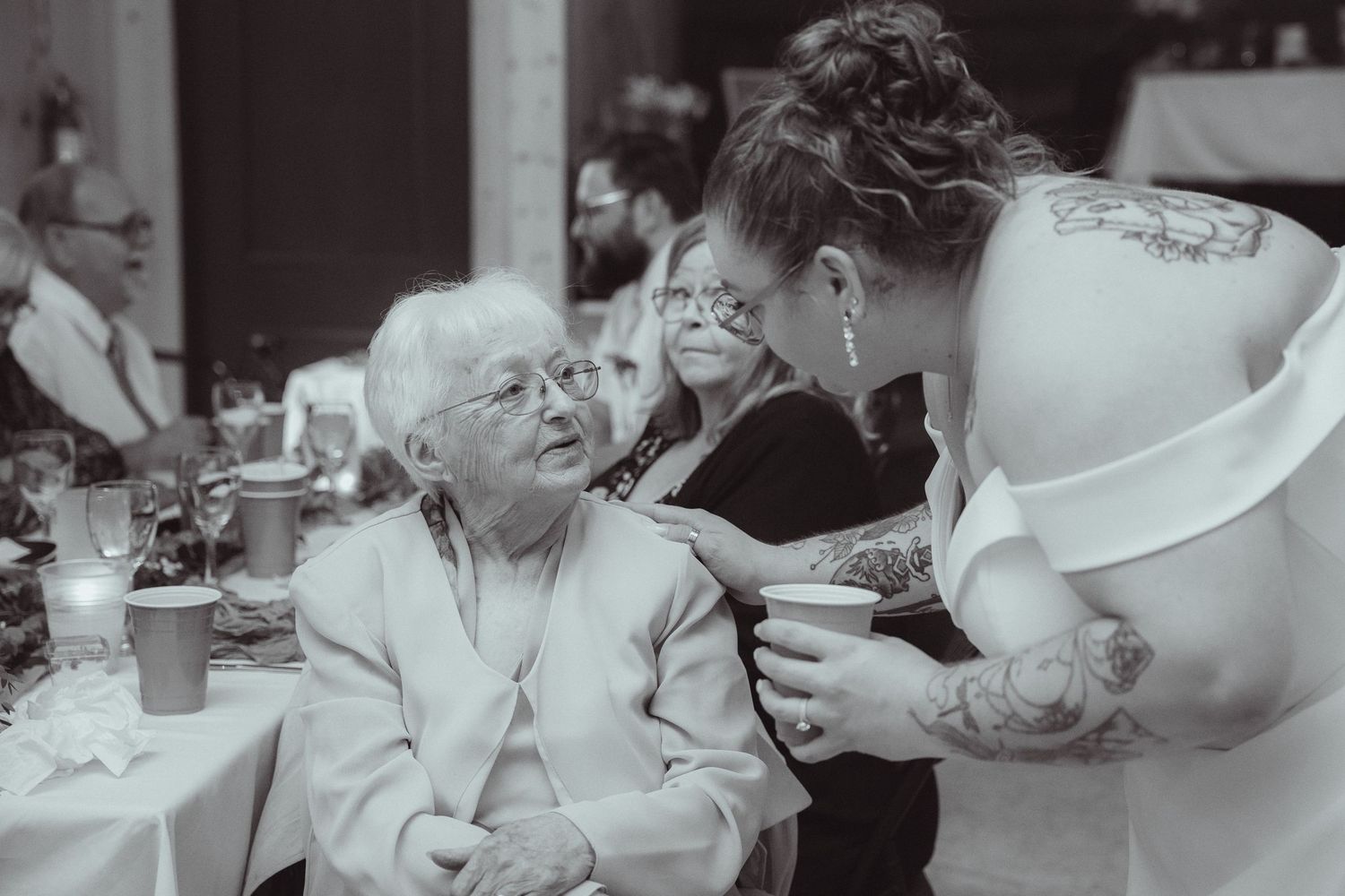 Black and white photo of tattooed bride leaning down to greet an elderly guest at her wedding reception.