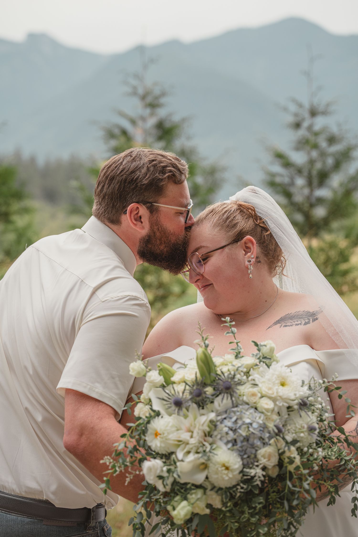 Groom kissing bride on forehead with mountain backdrop, bride holding white and blue bouquet wearing veil and glasses.