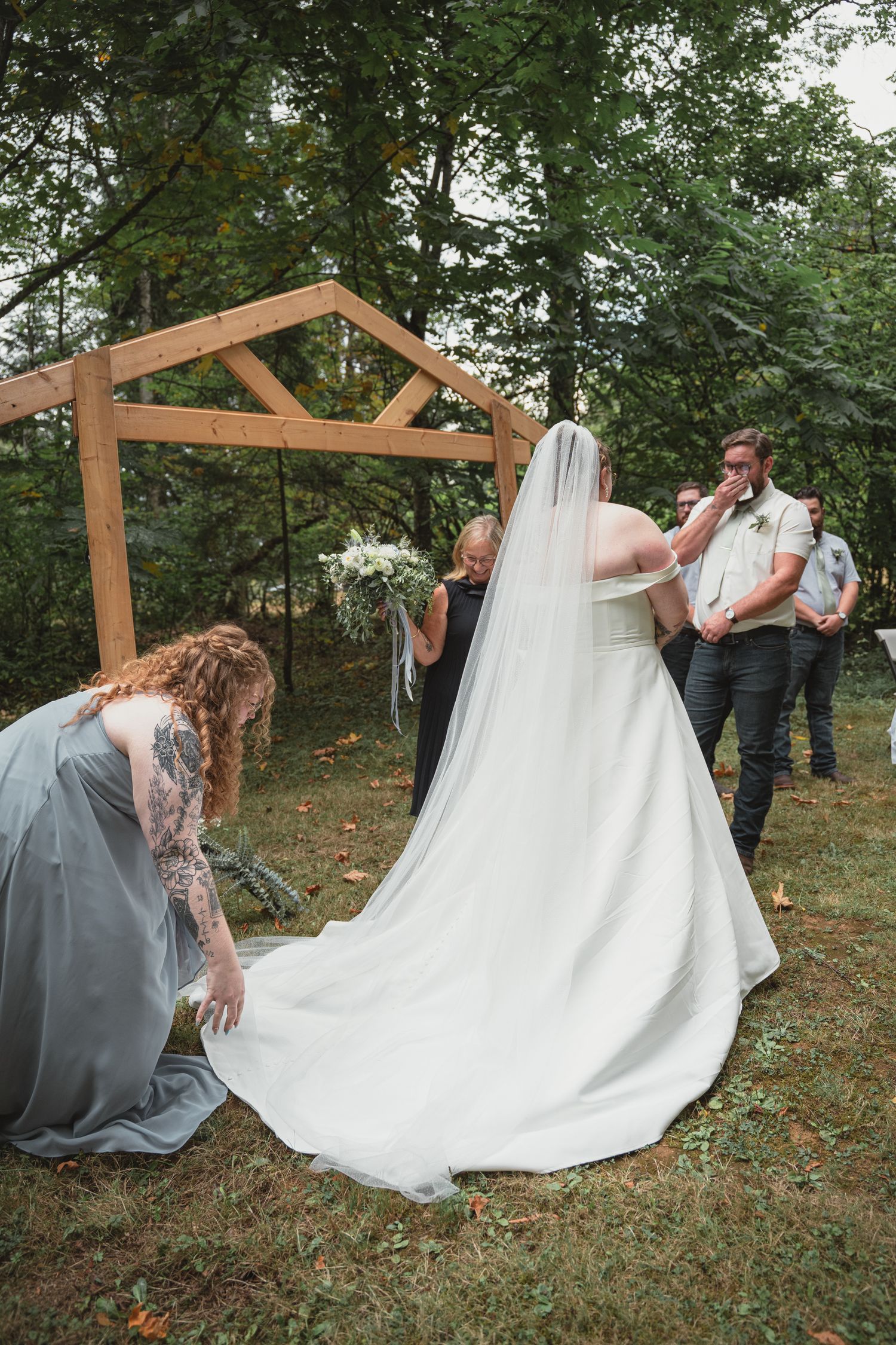 Bridesmaid adjusts bride's white wedding gown train at rustic wooden arch during outdoor forest ceremony.