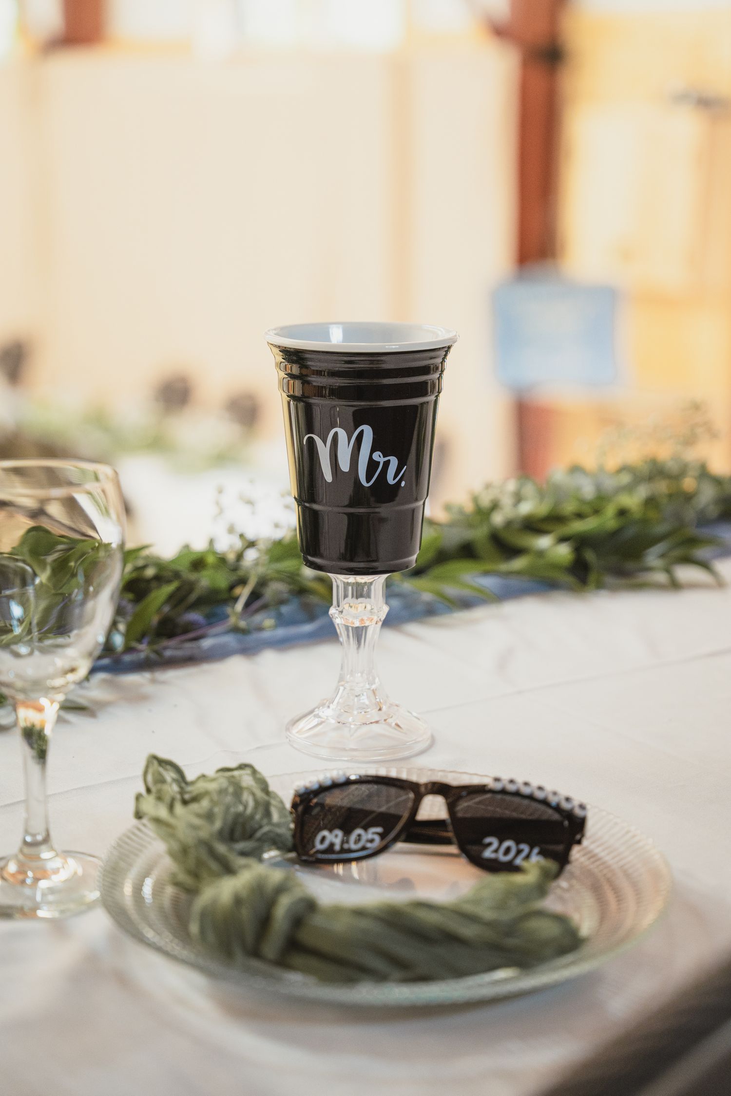 Wedding table setting with Mr. cup, sunglasses dated 09.05.2024, greenery, and glassware on white tablecloth.