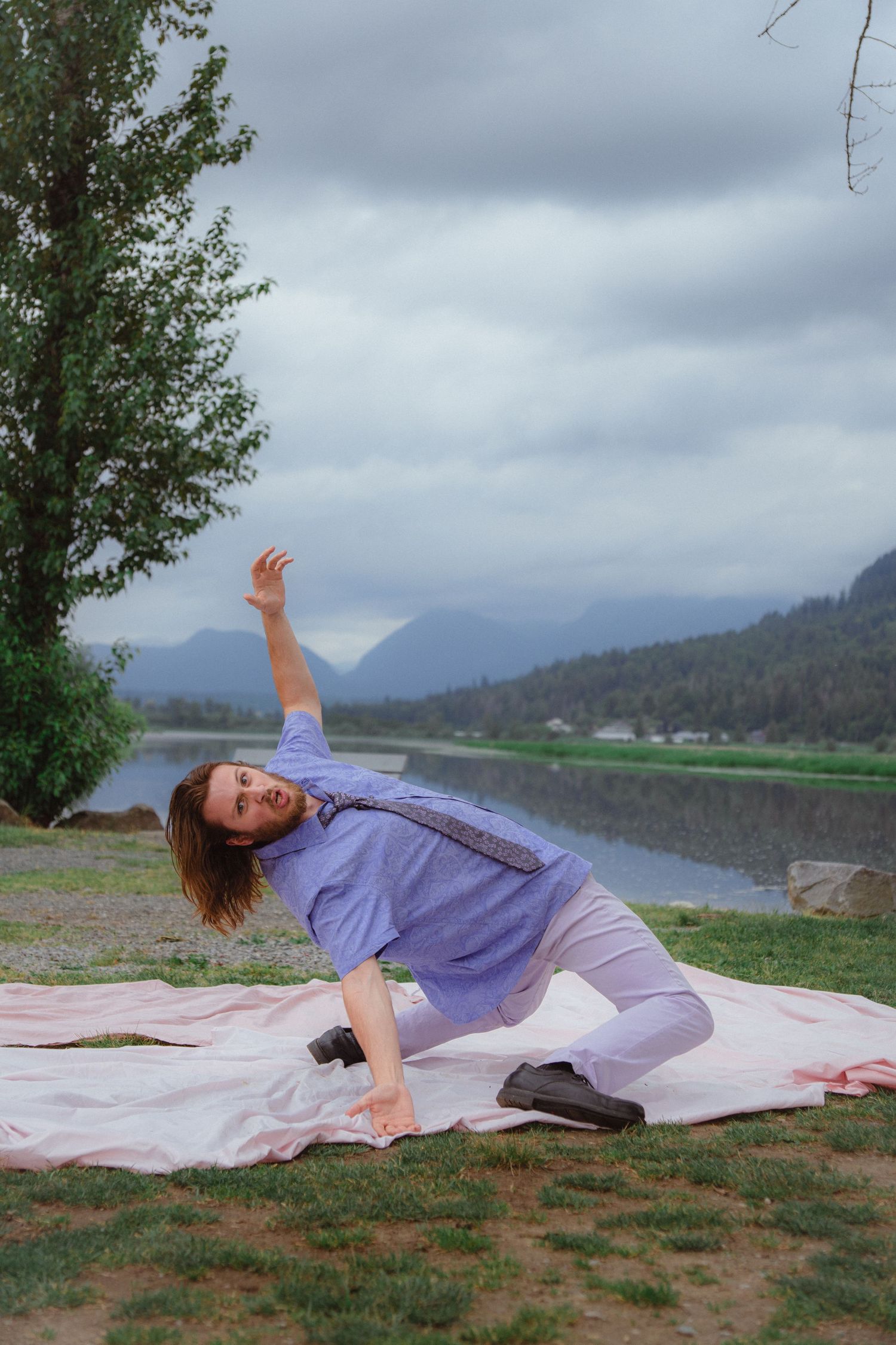 Man in blue shirt and gray pants poses dramatically on pink blanket by mountain lake.