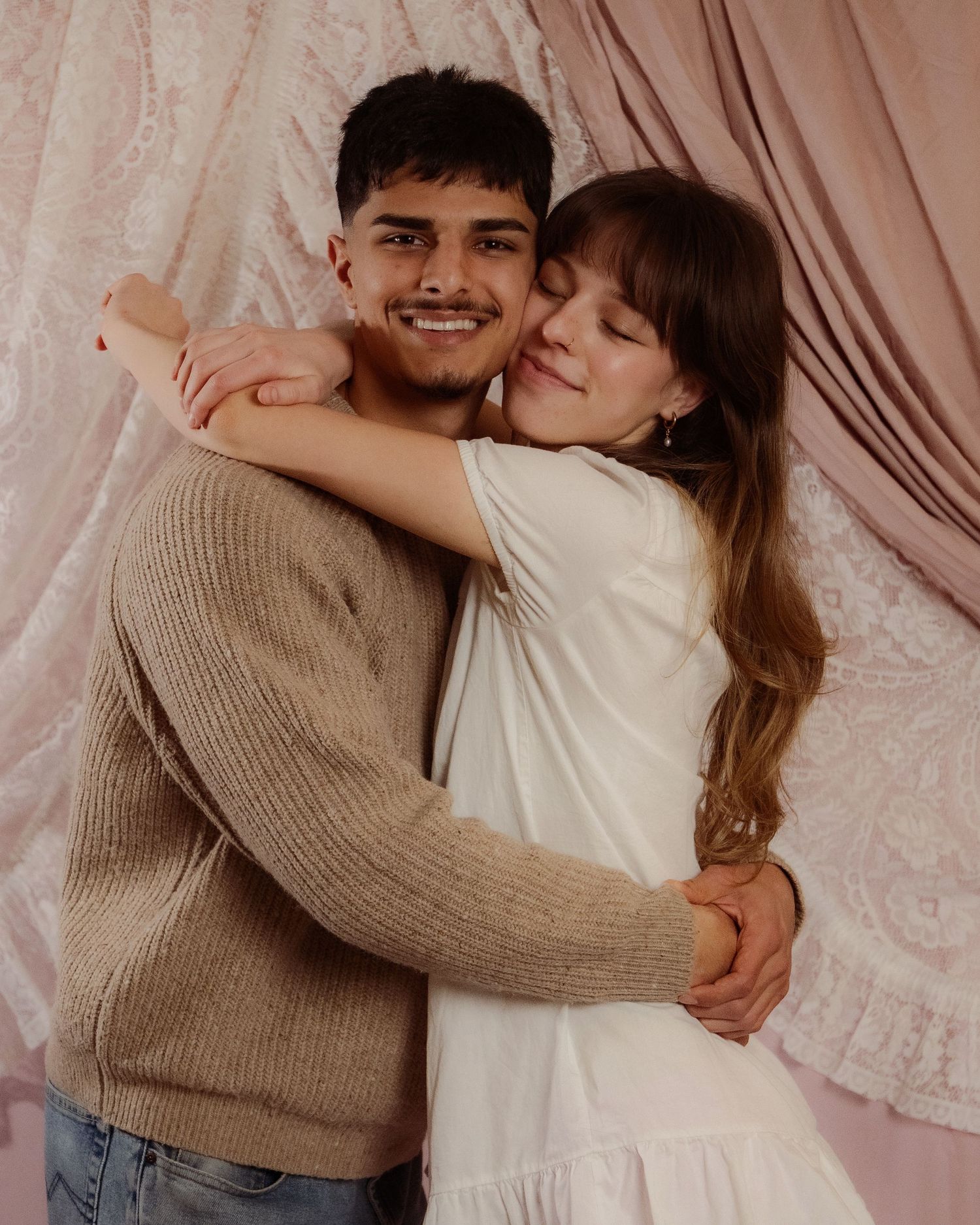 Young couple sharing a warm embrace, smiling, against a pink lace and fabric backdrop.