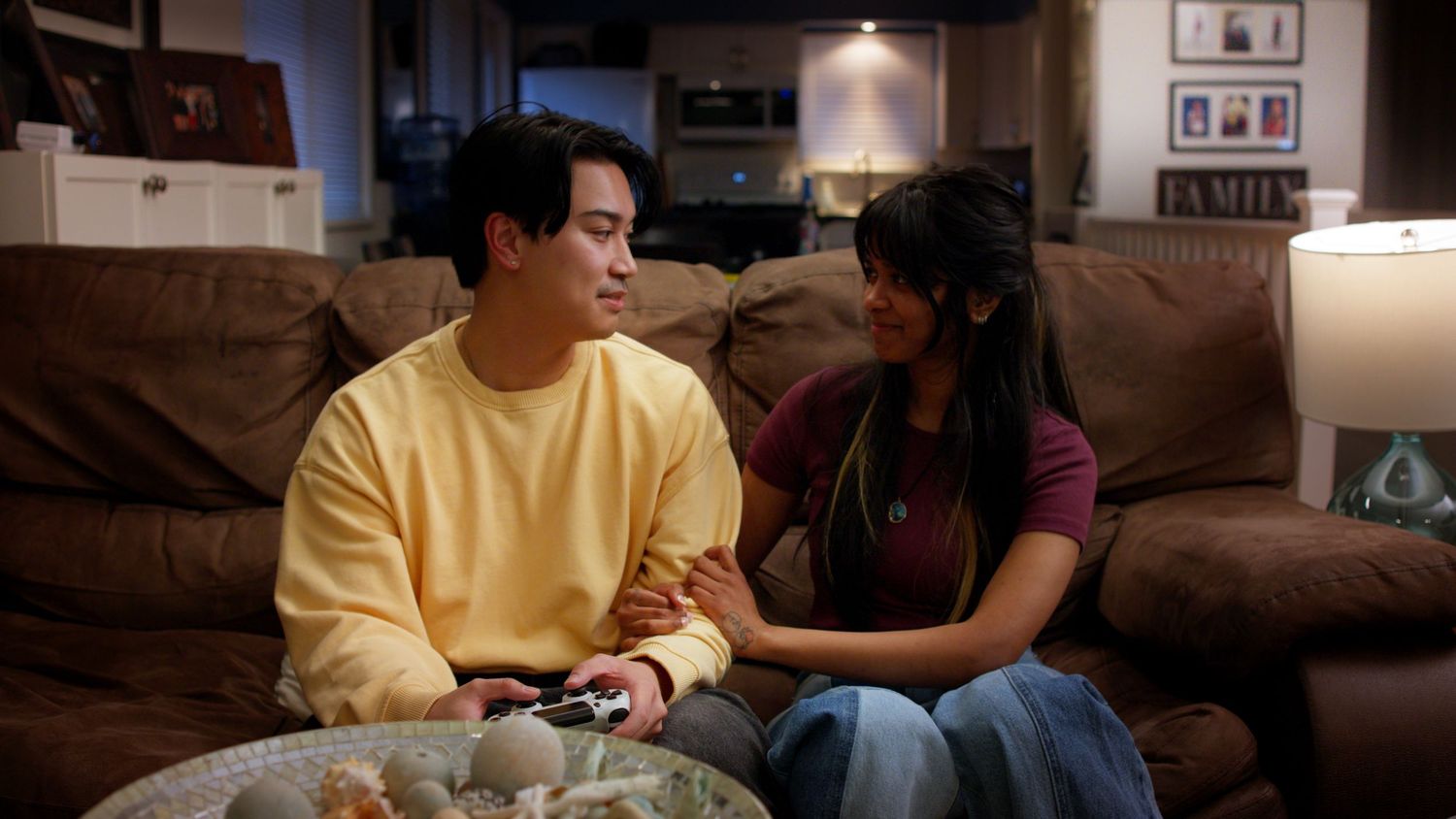 Young Asian man and woman sit on a couch together, with the woman placing her hand on his arm.