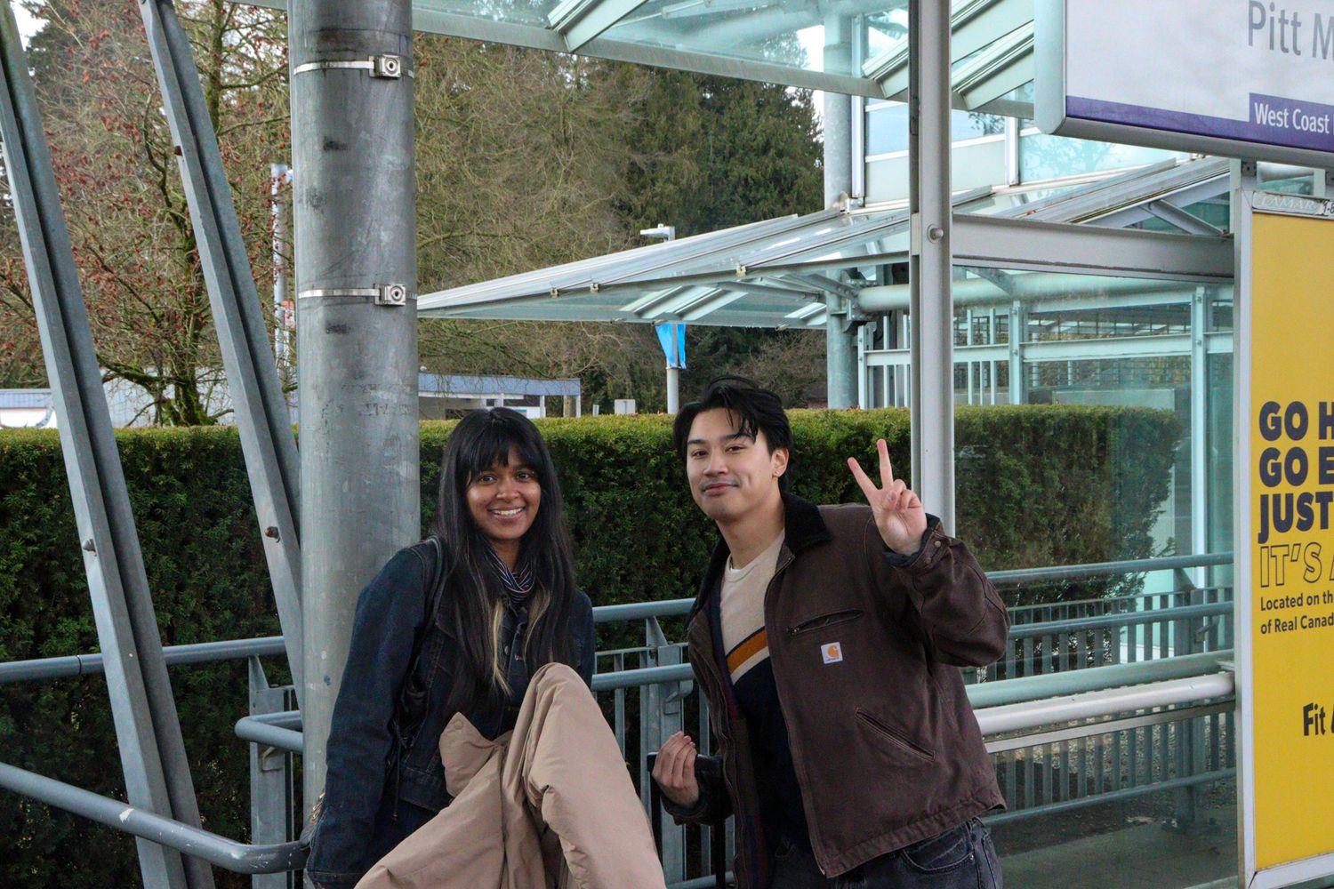 Two smiling people pose at Pitt Meadows SkyTrain station entrance.