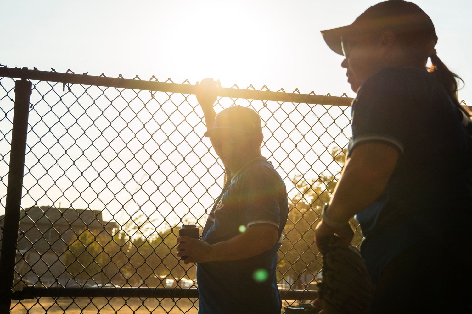 Two softball players silhouetted against bright sunlight near a chain-link fence at golden hour.