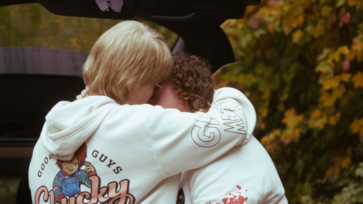 Two people hugging warmly while wearing matching white Chucky graphic hoodies near a car trunk in autumn.