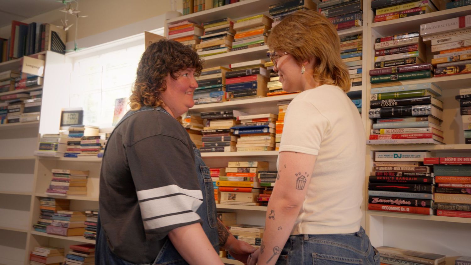 Two young women holding hands and smiling at each other in front of a bookshelf.
