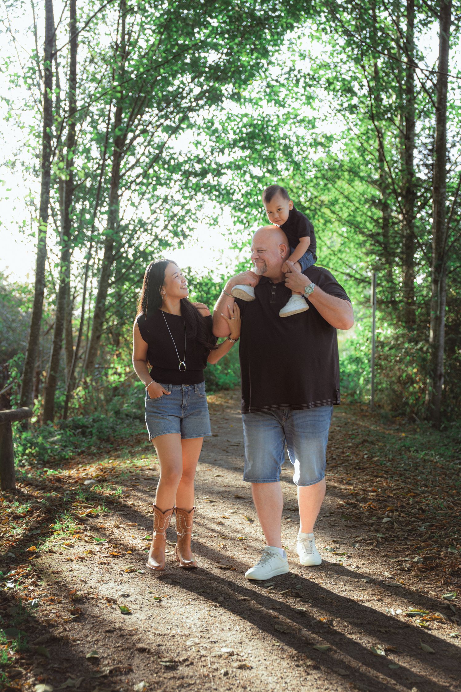 A happy family of three walks on a sunny forest path, with a child sitting on dad's shoulders.