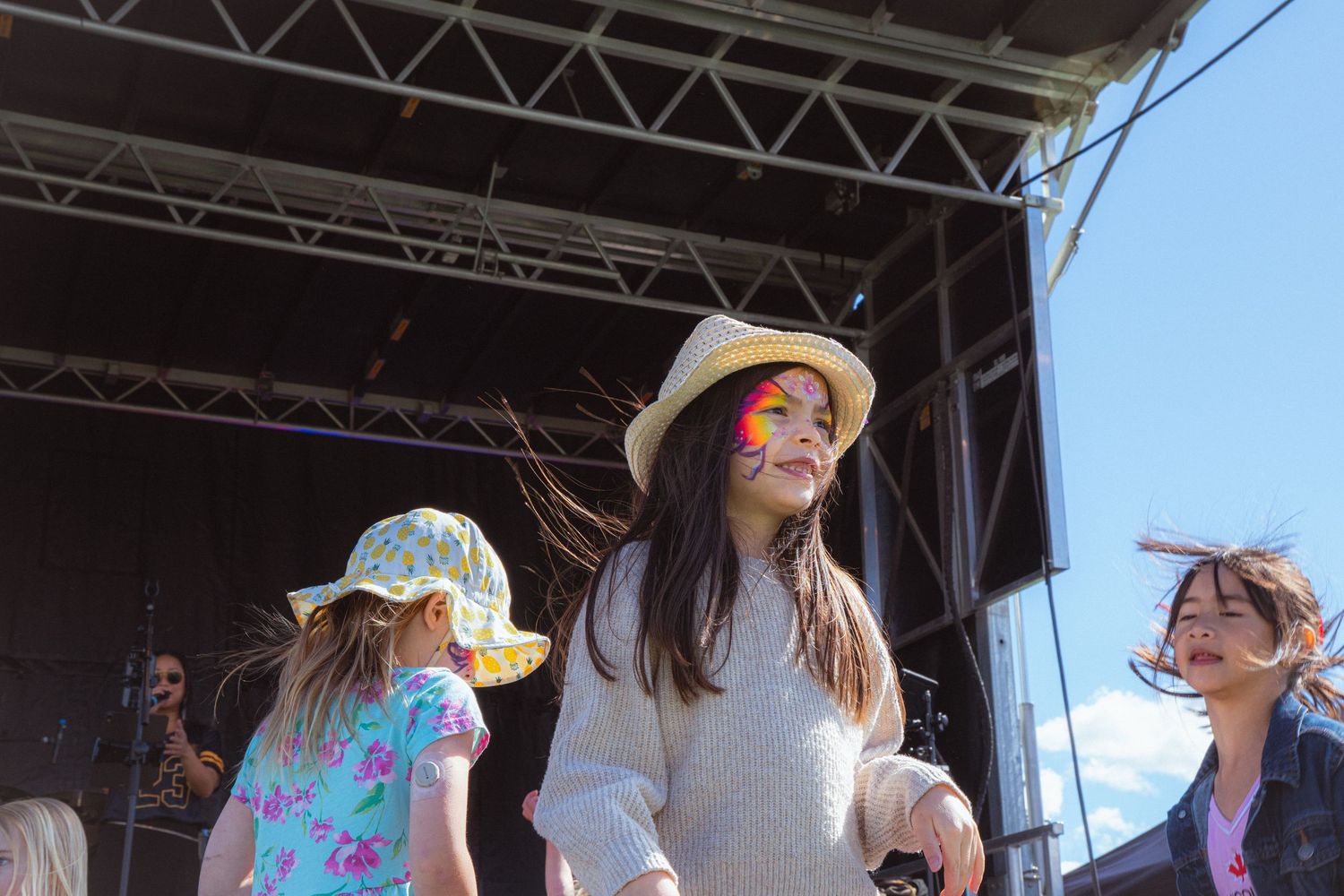 Young girls with face paint dancing near outdoor concert stage on sunny day.