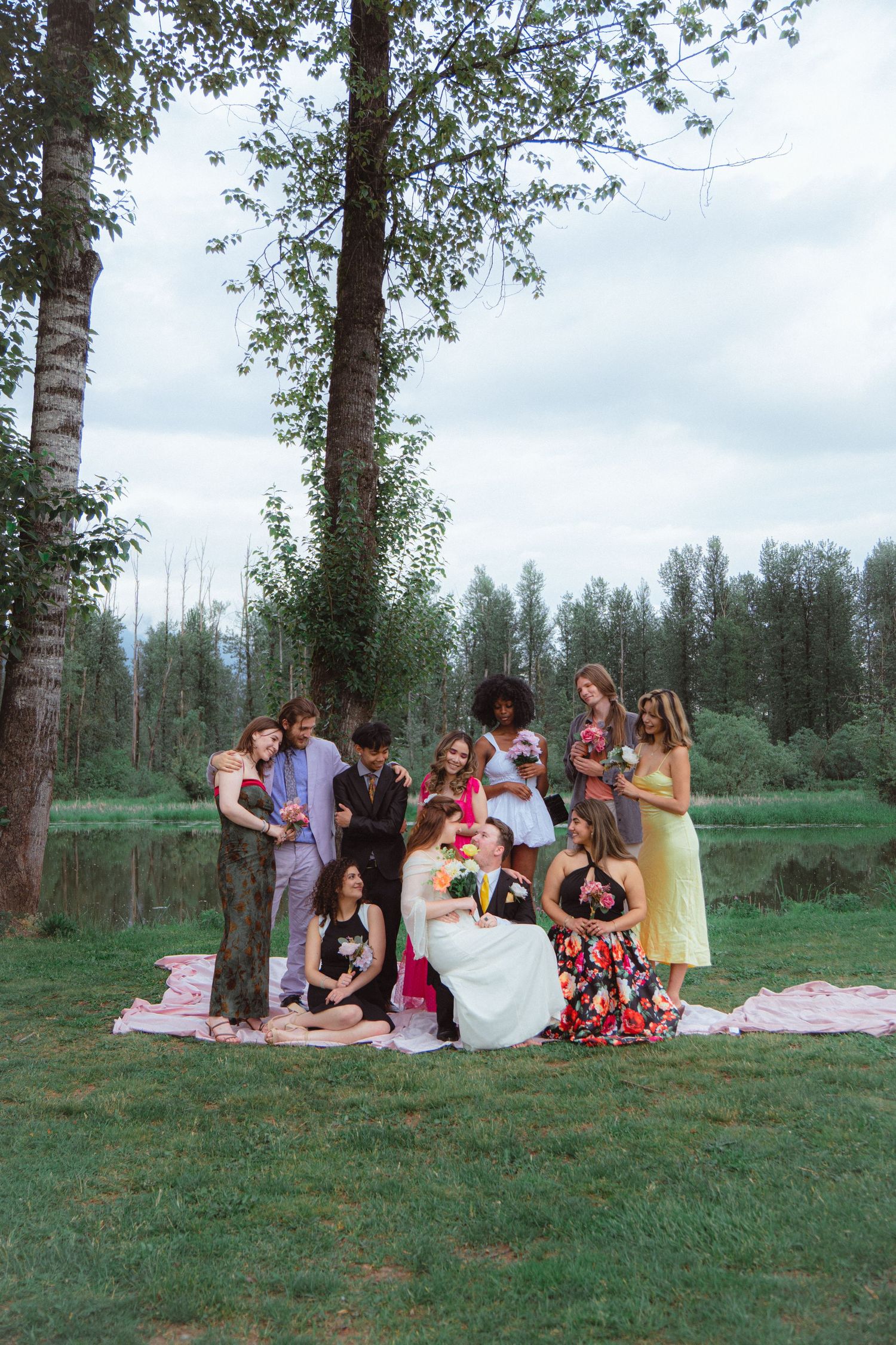 Wedding party gathers around bride and groom for a candid lakeside group photo.