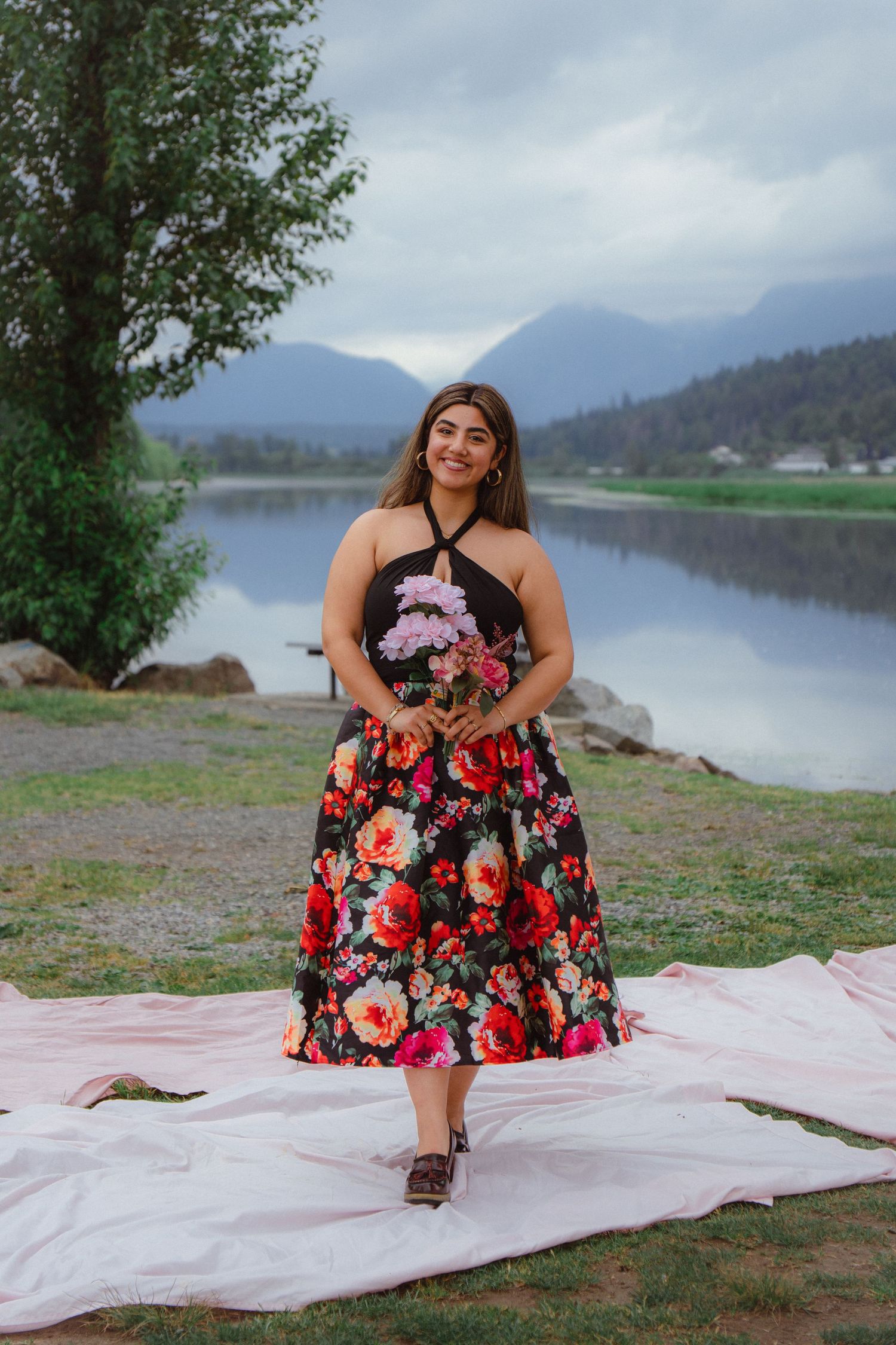 Woman in floral midi dress holding pink flowers standing by a calm reflective mountain lake.