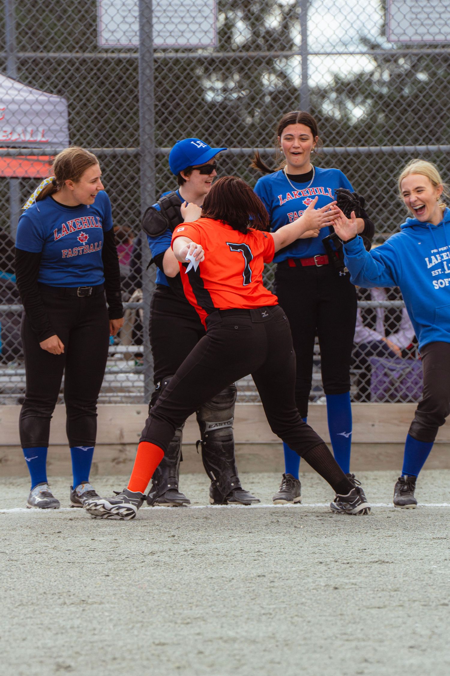 Softball players in blue Lakeville uniforms celebrate with opponent number 7 in orange jersey on the field.