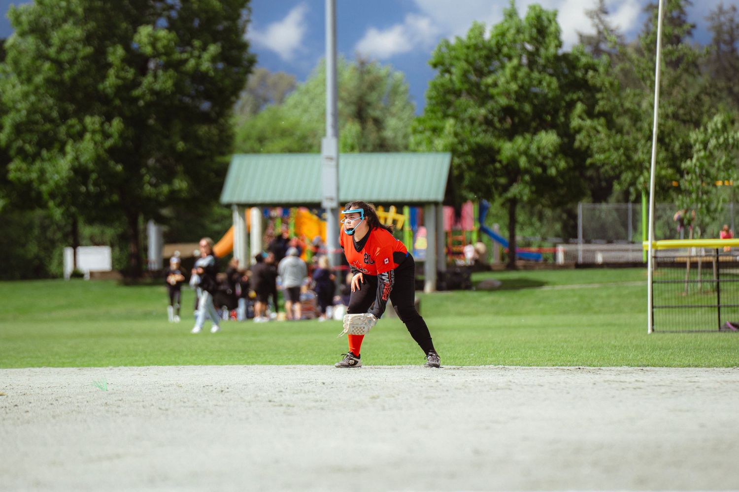 Softball player in orange uniform crouches at base on sunny outdoor field near playground area.