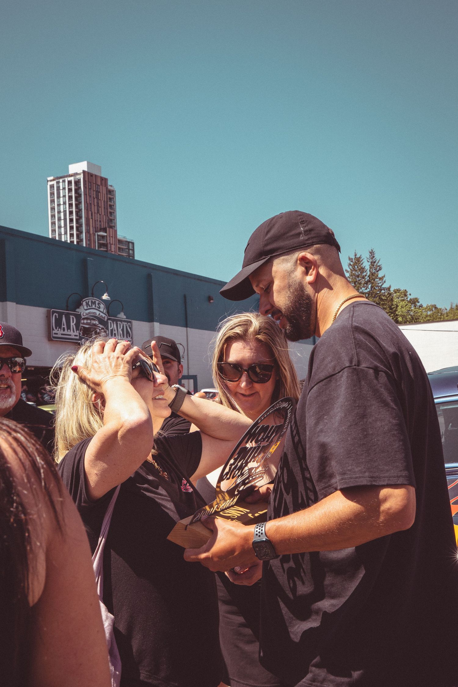 Man in black cap showing trophy to two women wearing sunglasses at outdoor car show event.