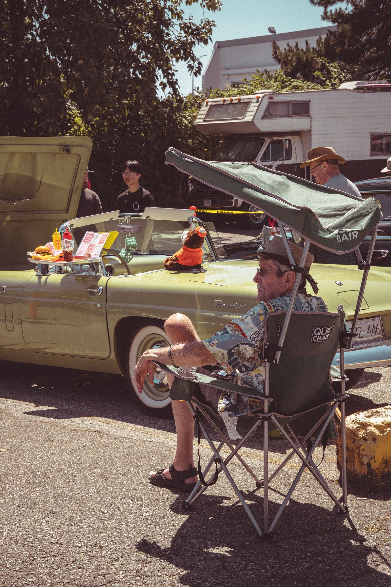 Man relaxing in camping chair beside a vintage mint green convertible car at an outdoor classic car show.