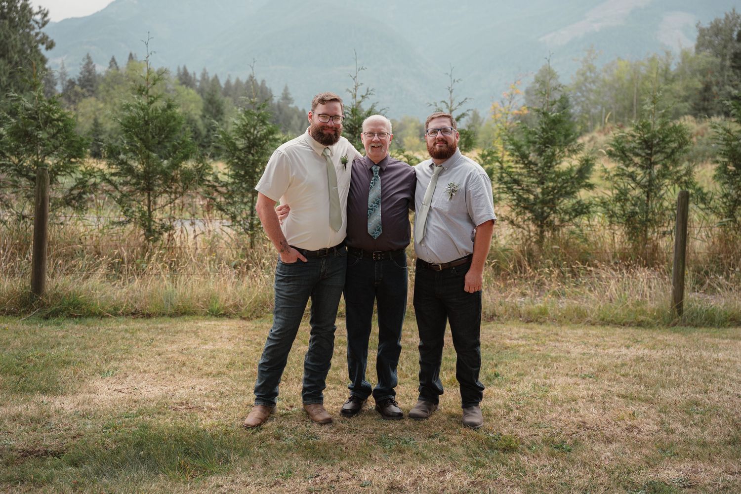 Three men smiling together outdoors at a wedding with mountain scenery behind them.