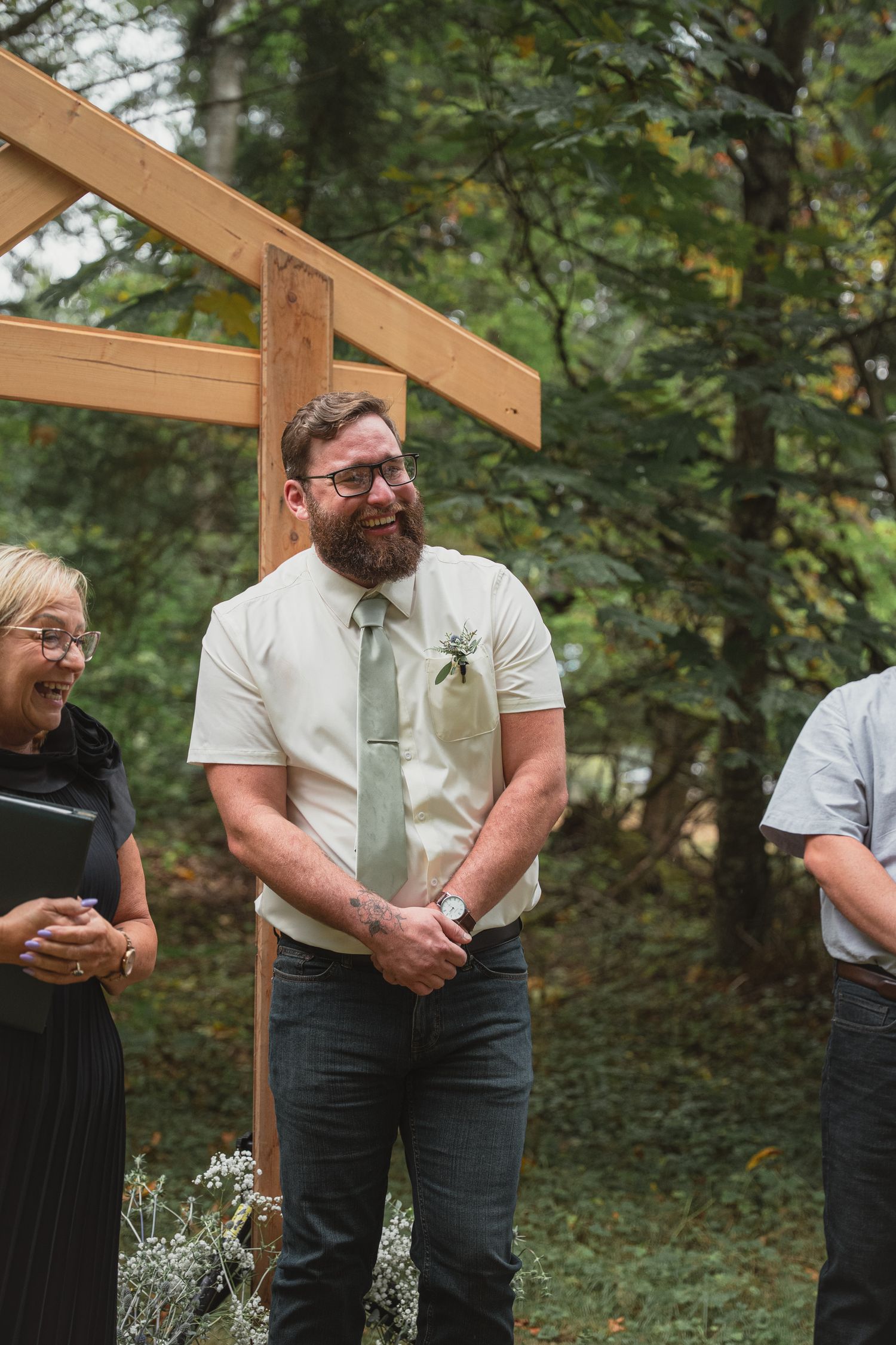 Bearded groom in white shirt and sage tie smiling joyfully during outdoor wedding ceremony in wooded setting.