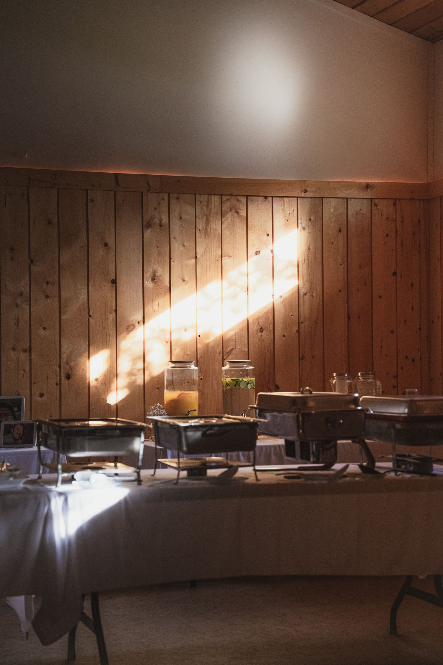 Buffet table with chafing dishes and drink dispensers lit by sunlight against wood panel wall.