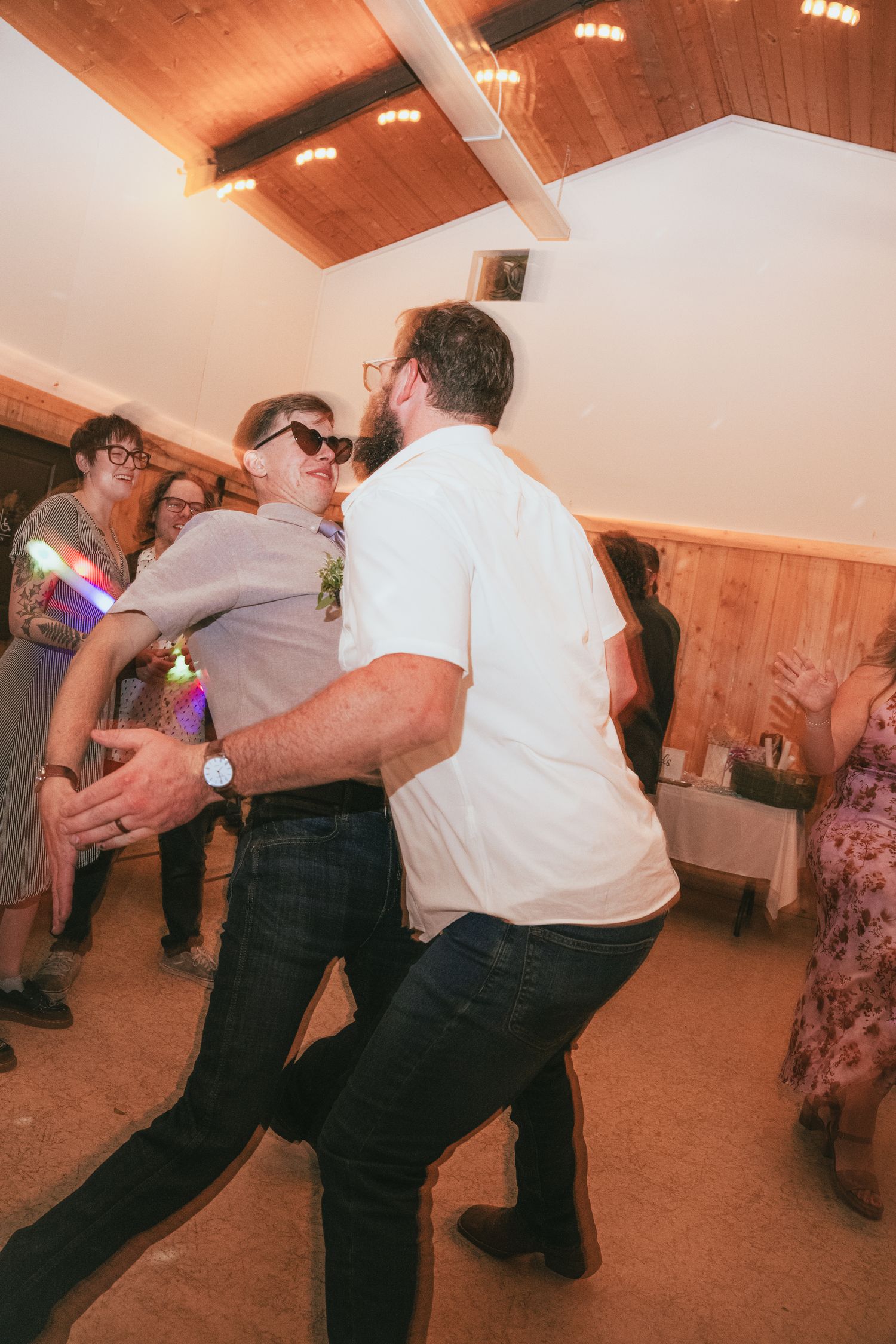 Two men dancing energetically at a wedding reception in a wood-paneled venue with guests celebrating in the background.