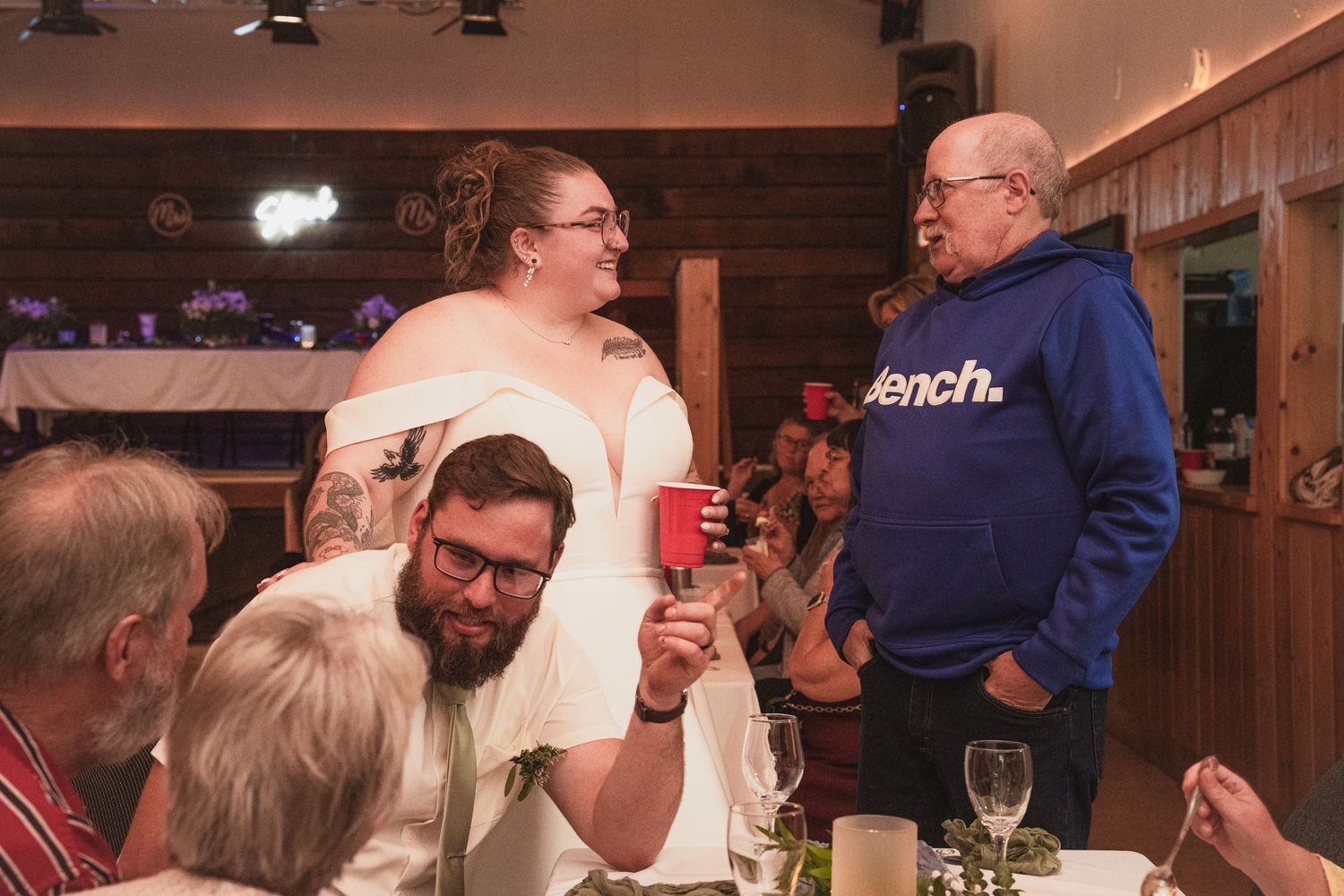 Bride and groom toast with red cups while greeting an older guest in a blue hoodie at their wedding reception.