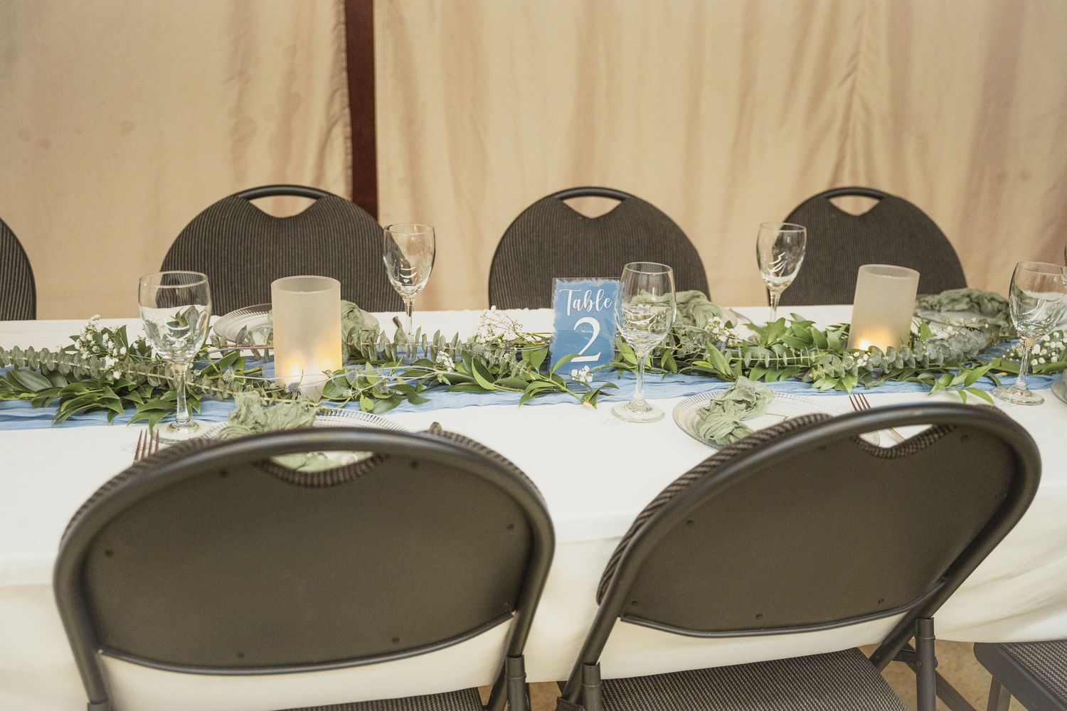 Elegant wedding head table with white linens, greenery garland, candles, and a blue table number card in a tent venue.