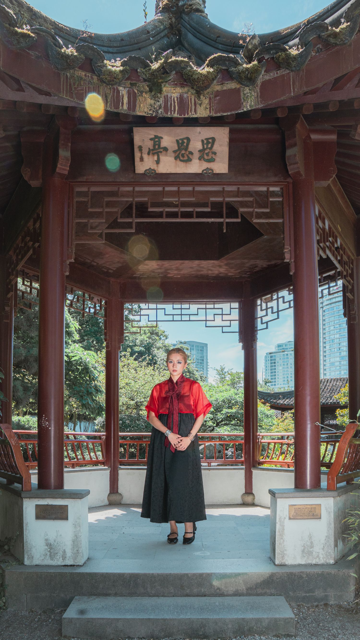 Woman in red top and black skirt standing in traditional Chinese garden pavilion.