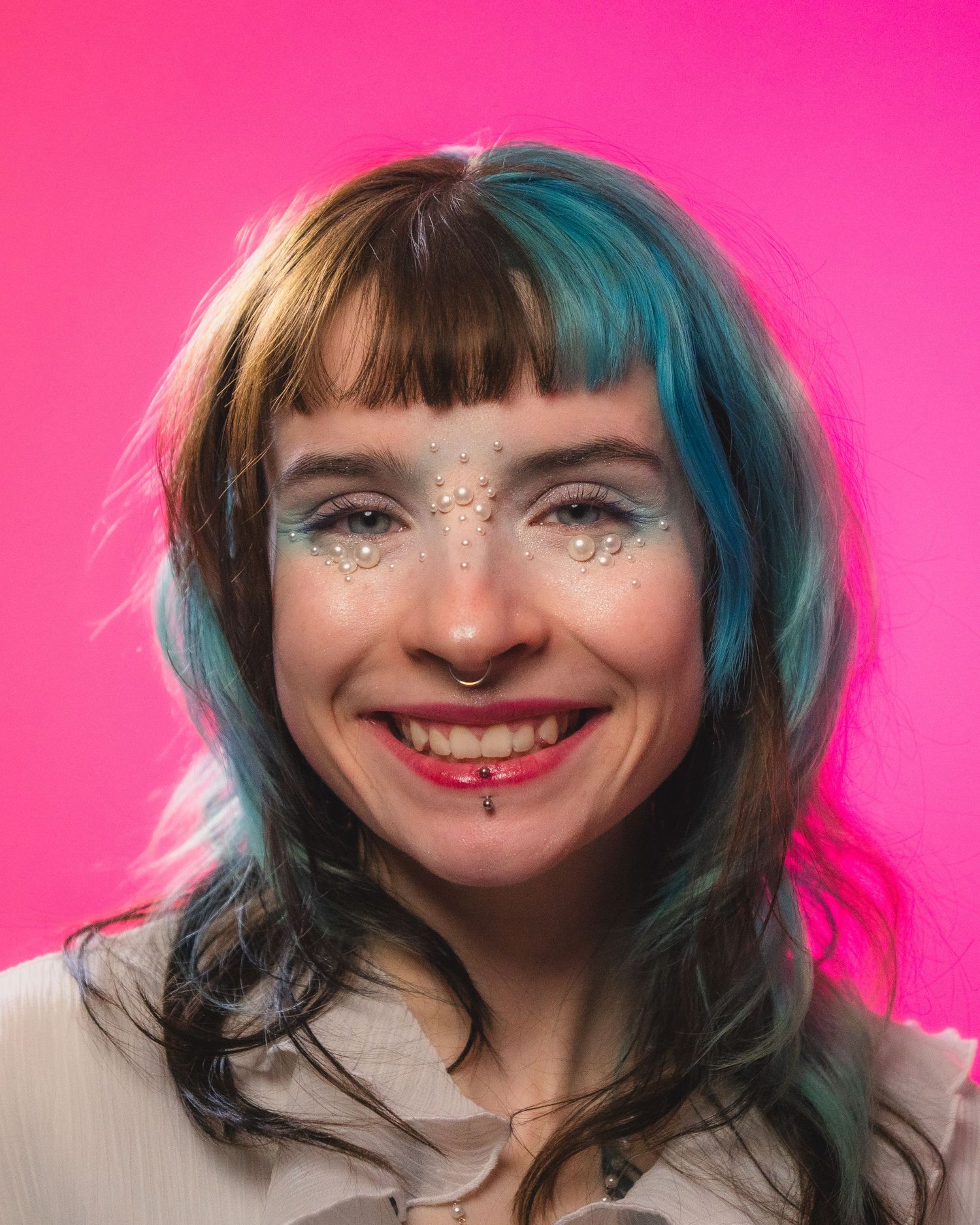 Smiling woman with teal hair, nose ring, and glitter face gems on vibrant pink background.