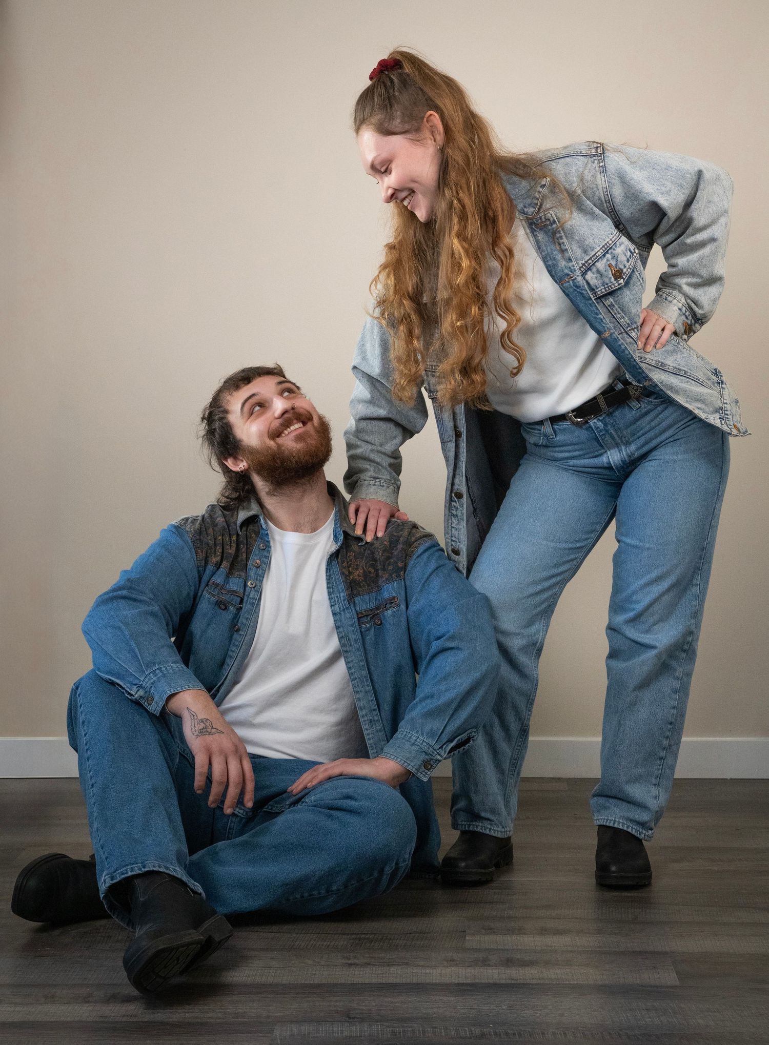 A smiling couple in matching denim outfits share a joyful moment, man seated on floor woman standing beside him.