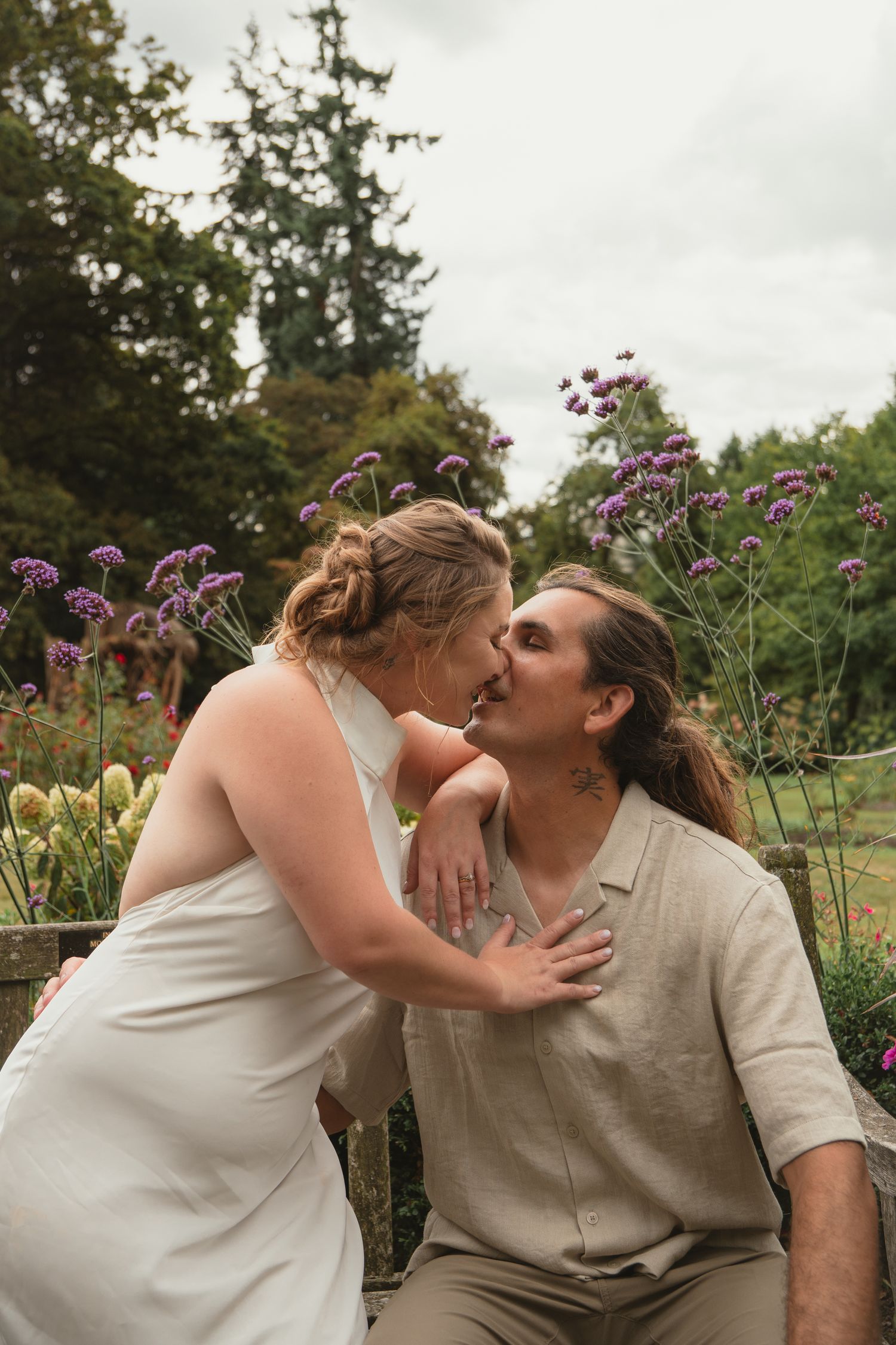 Bride and groom share a tender kiss seated among purple wildflowers in a lush garden setting.
