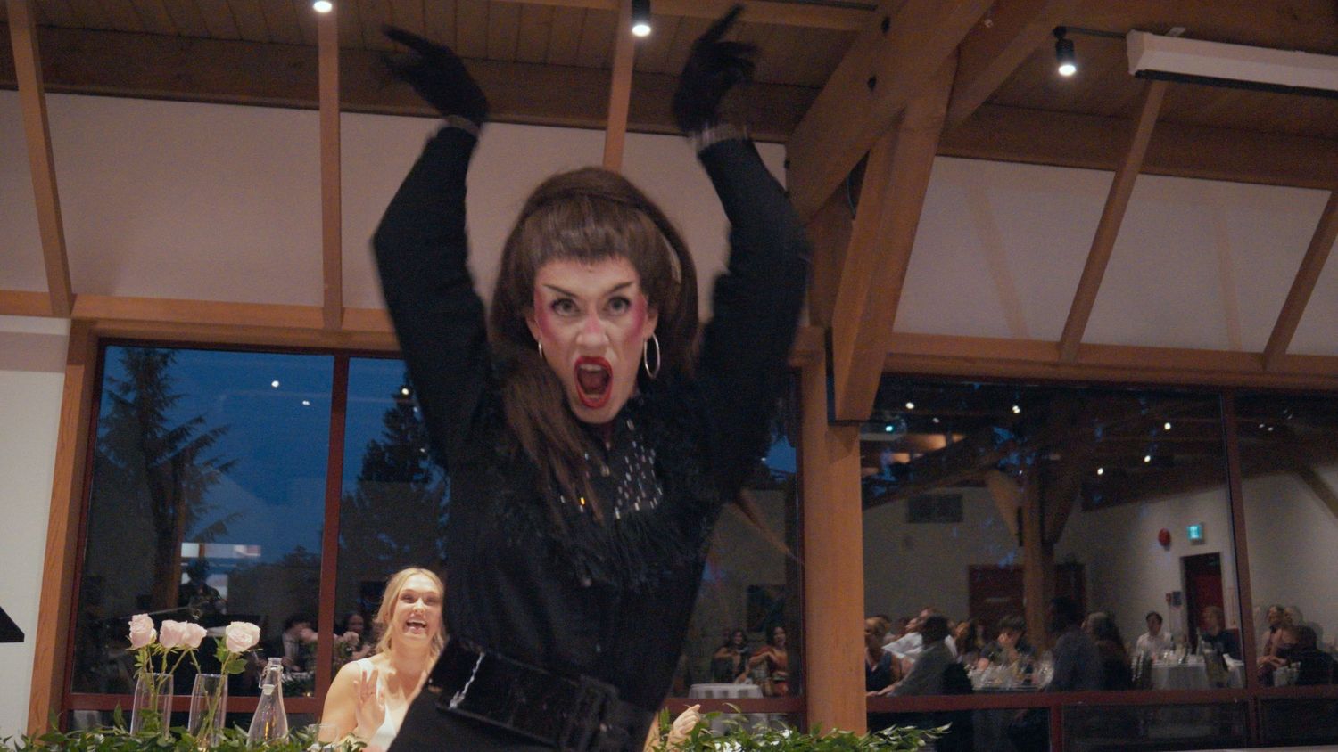 Drag performer in black outfit dramatically posing at a wedding reception venue with guests in background.