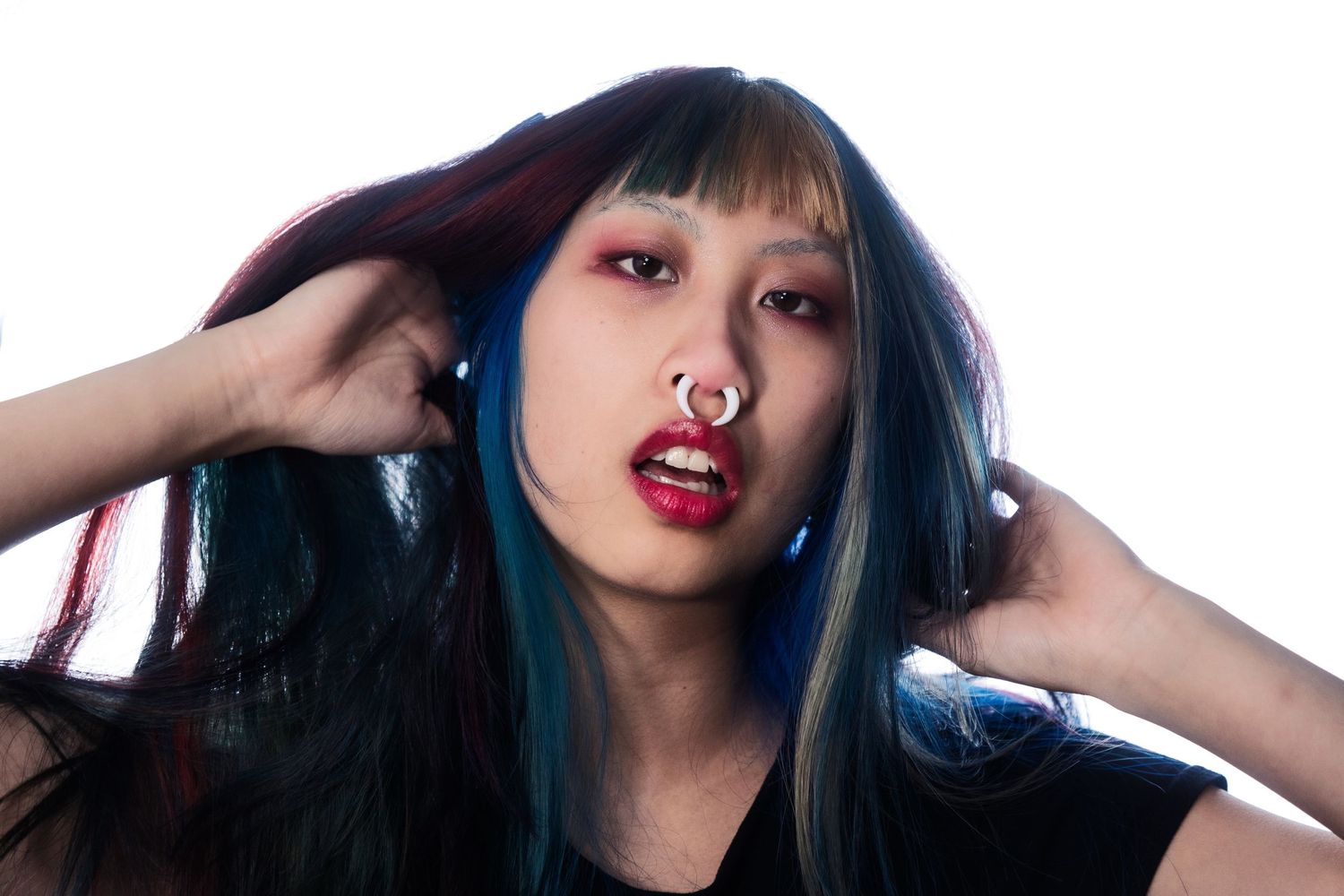 Young woman with vibrant blue and red streaked hair, septum piercing, and bold red lips posing.