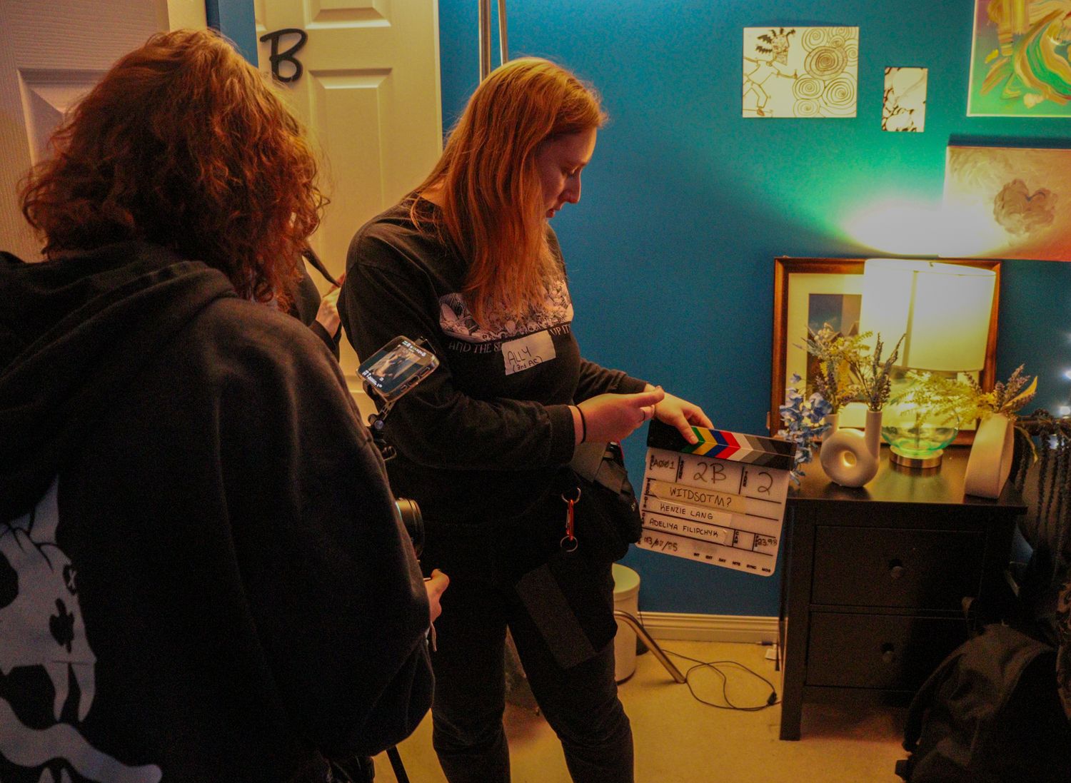 Two women examine a film clapperboard on a movie set with teal walls and warm lighting.