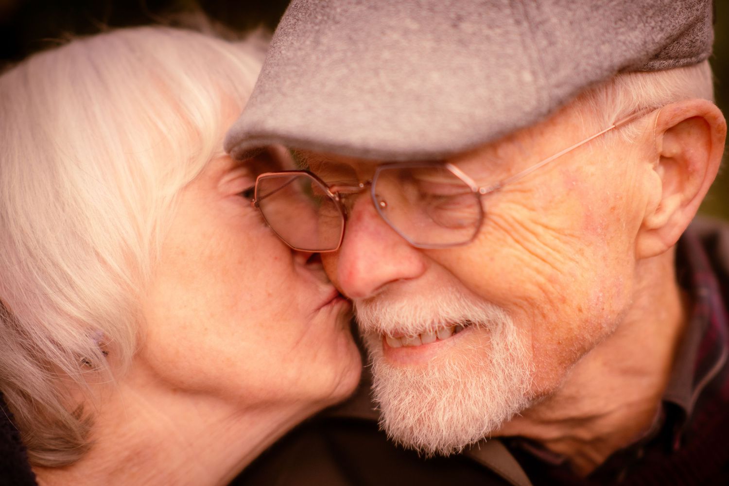 Elderly woman kissing senior man on the cheek as he smiles warmly outdoors.