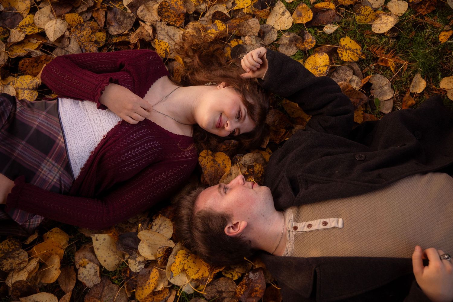 Couple lying in autumn leaves, woman in burgundy sweater and man in dark jacket, gazing at each other outdoors.