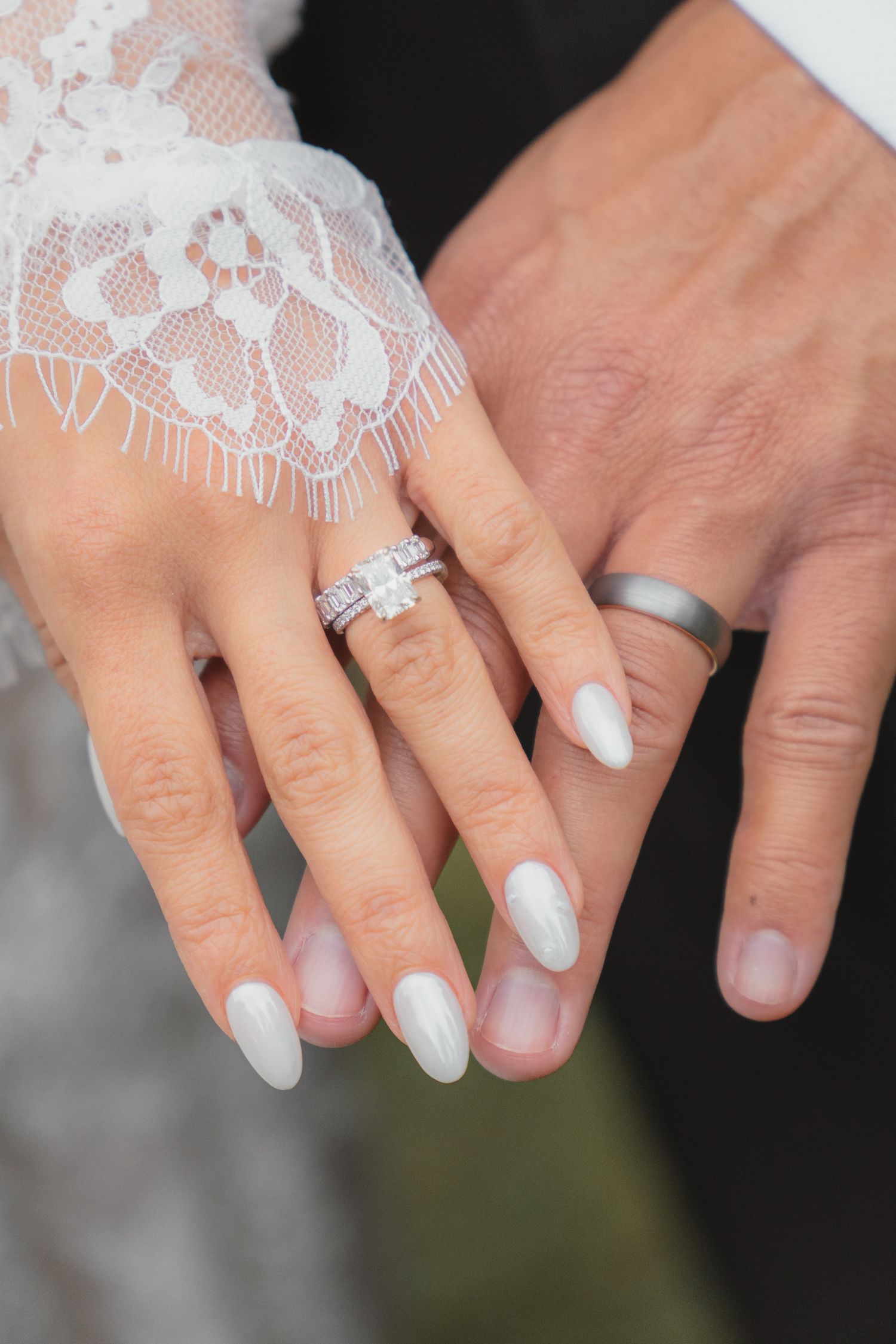 Newlyweds displaying wedding rings, bride wearing diamond ring with white manicure, groom with silver band, lace sleeve visible.