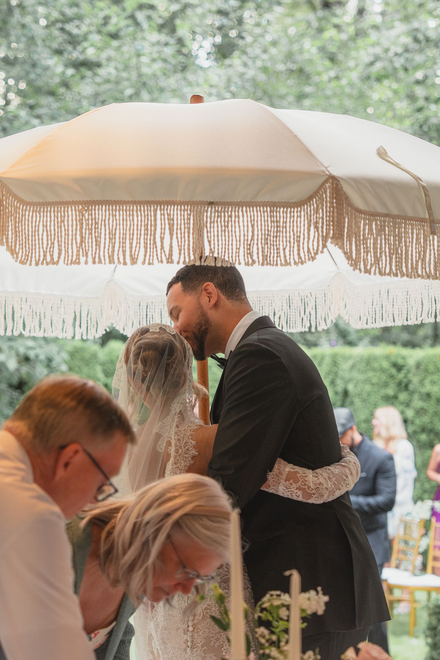 Couple sharing first kiss at outdoor wedding ceremony under fringed umbrella canopy surrounded by guests.