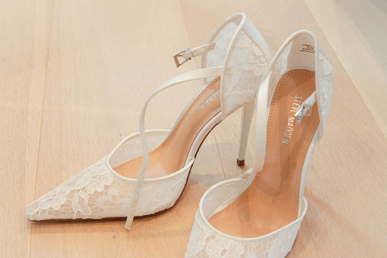 Elegant white lace bridal heels with ankle strap and stiletto heel on wooden floor.