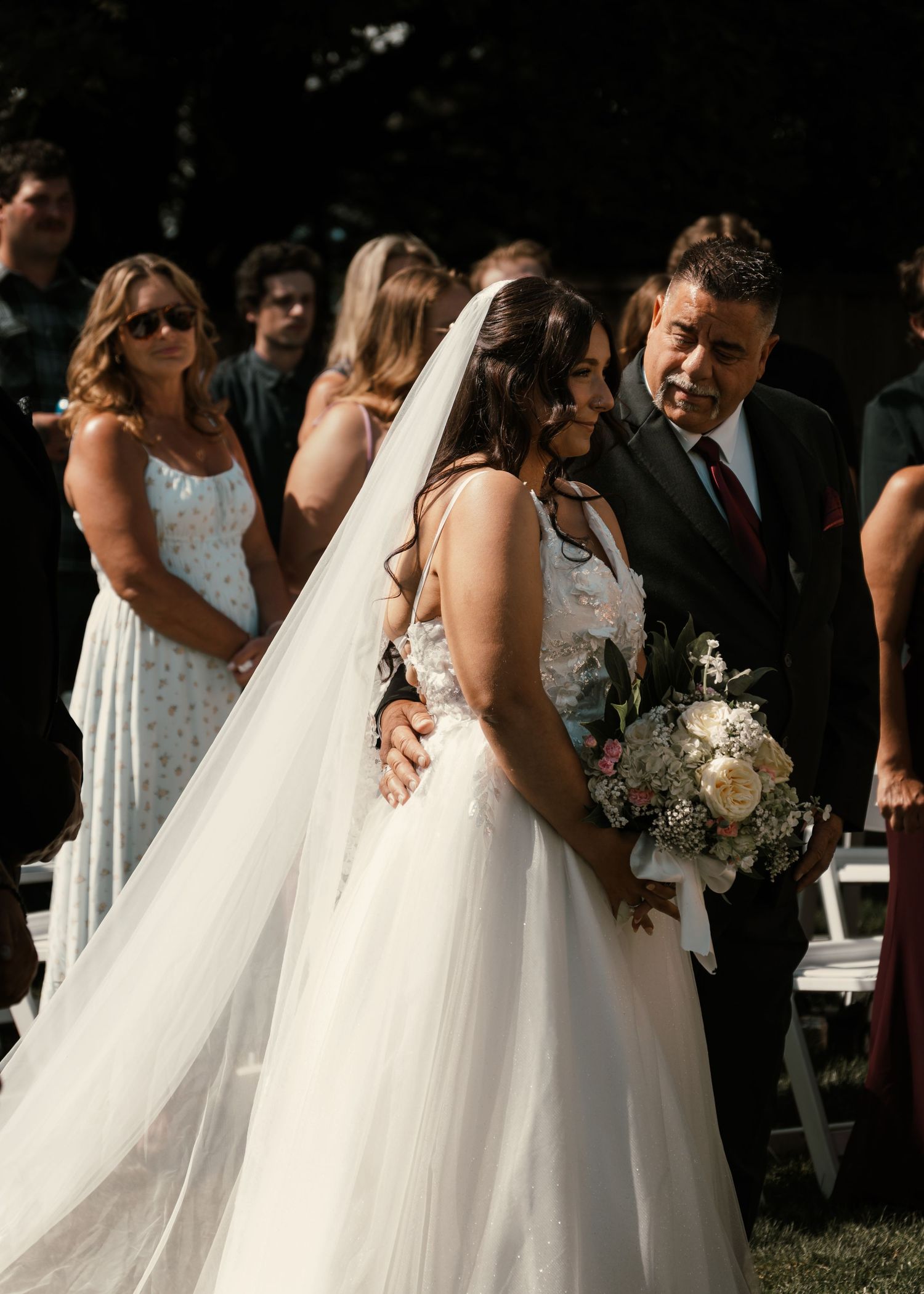 Bride in white gown and veil walks down outdoor aisle with escort, holding floral bouquet, guests seated in background.