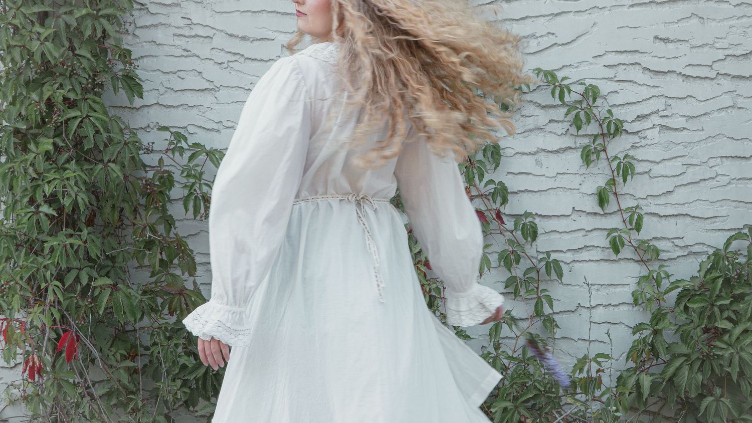 Woman in flowing white dress with long sleeves twirling outdoors against a stone wall with green ivy.