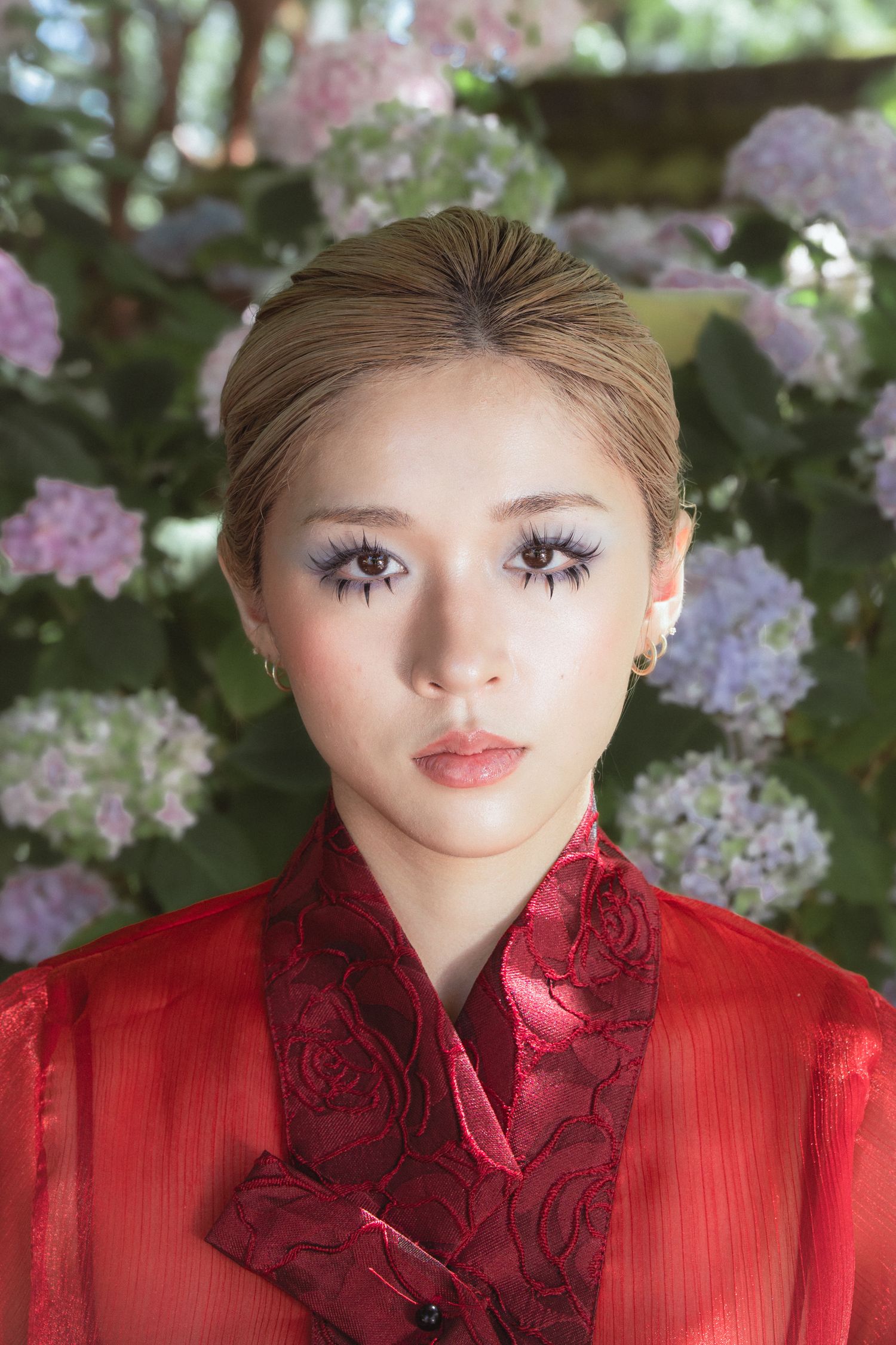 Young Asian woman in red floral qipao with dramatic lash makeup poses before blooming hydrangeas.