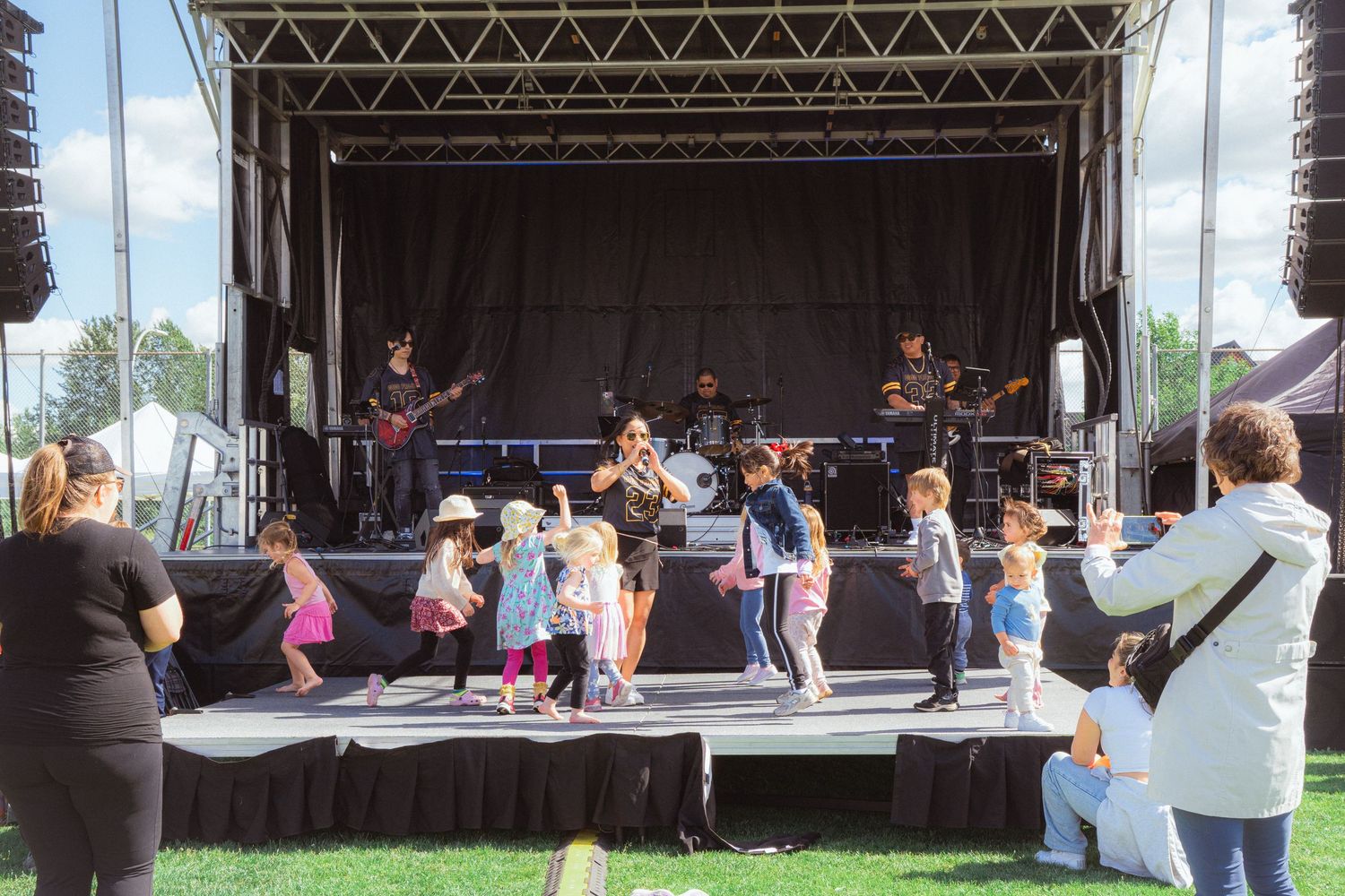 Band performing on outdoor stage while children dance in front with parents watching.