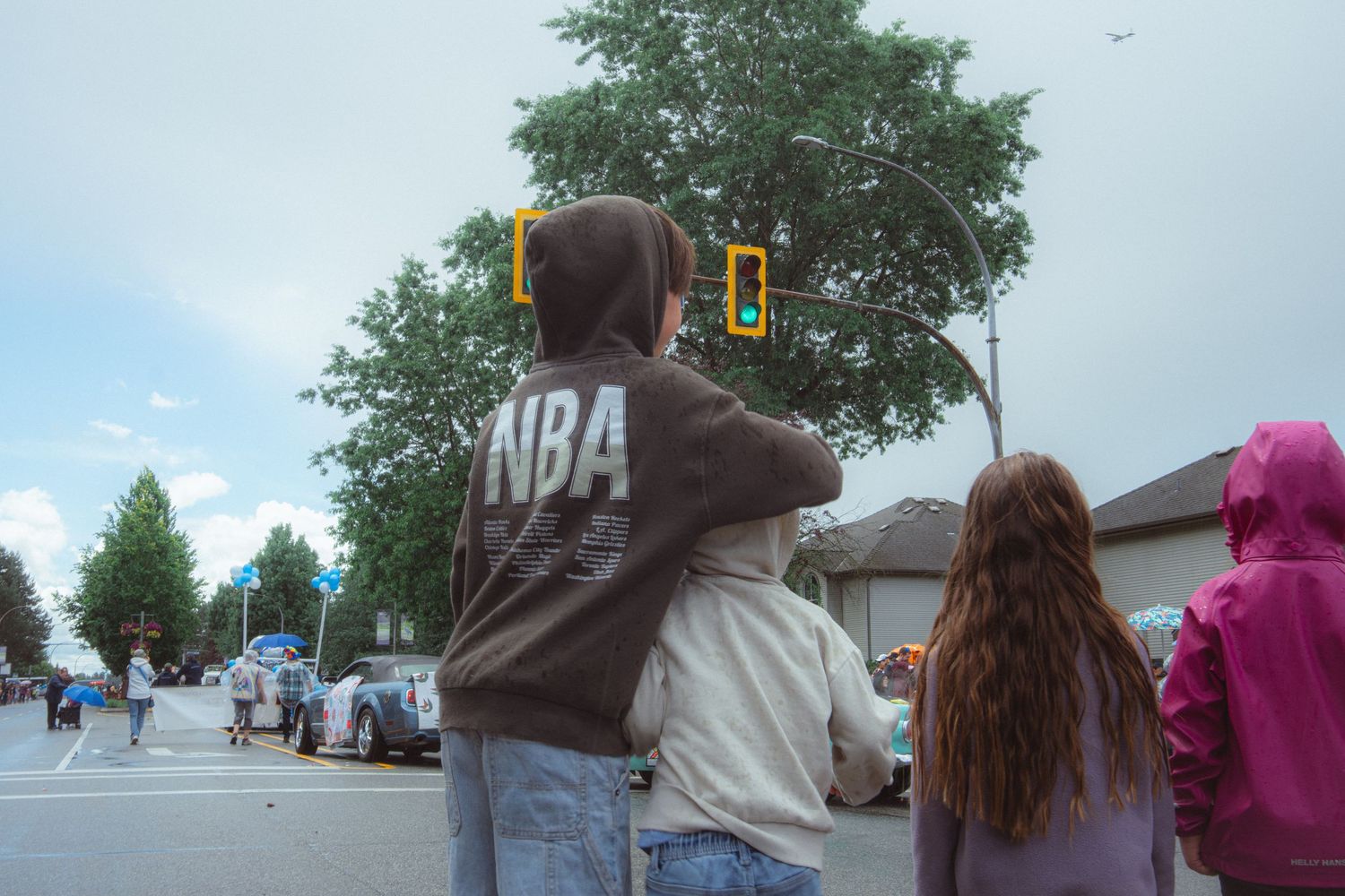 Children in NBA hoodies watch a street parade from the sidewalk on an overcast day.