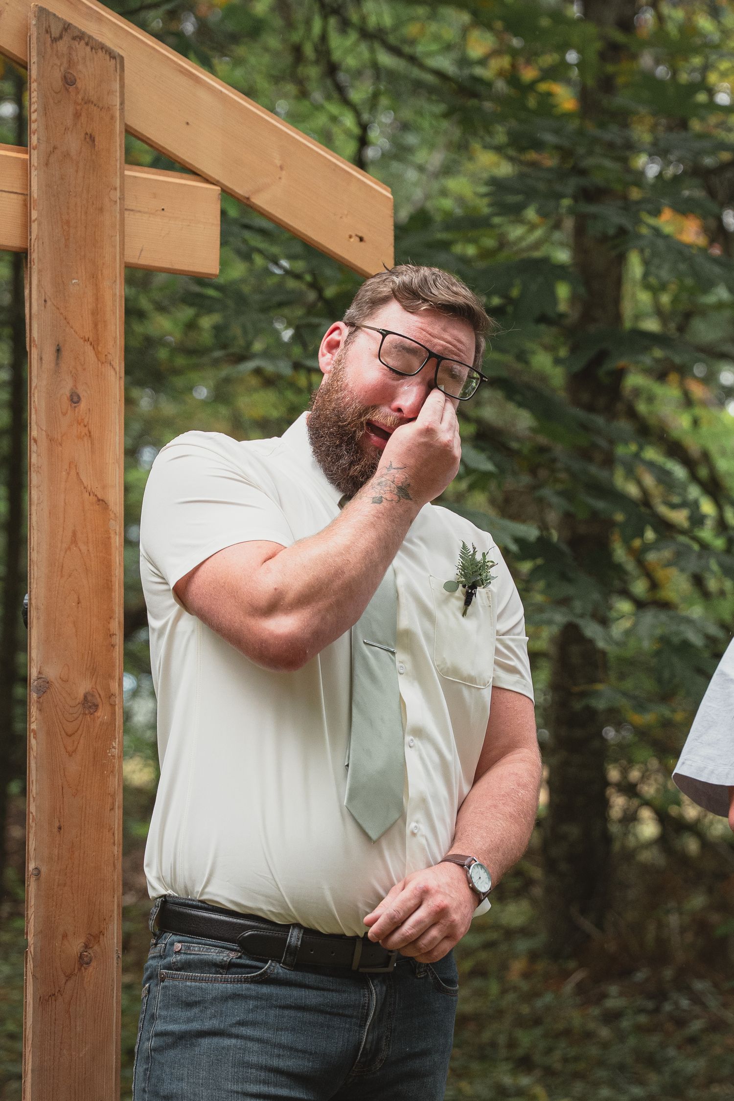 Emotional groom wiping tears at outdoor wedding ceremony, wearing glasses, white shirt, and sage green tie with boutonniere.