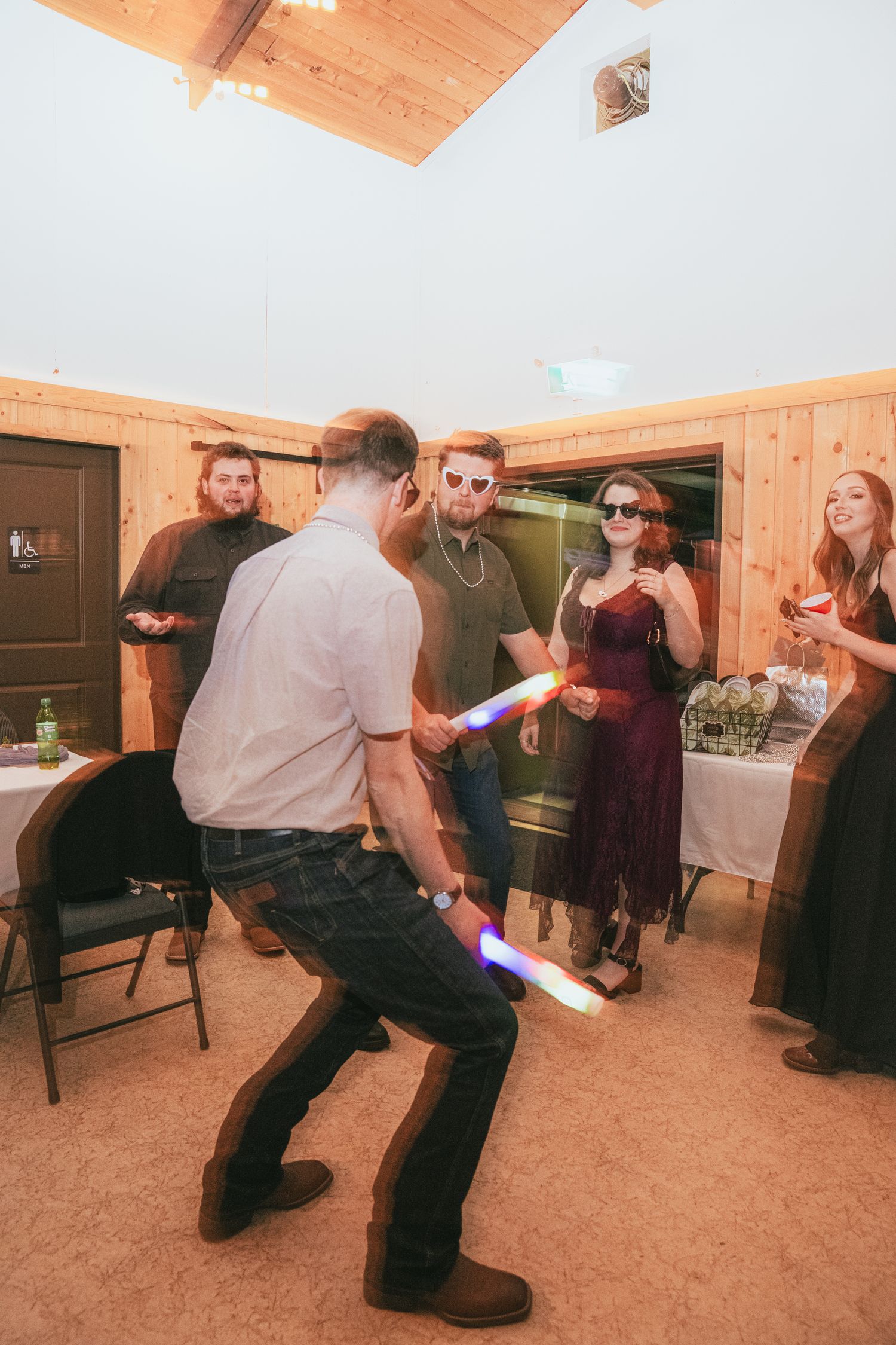 Guests dancing with colorful glow sticks at rustic wooden wedding venue reception.