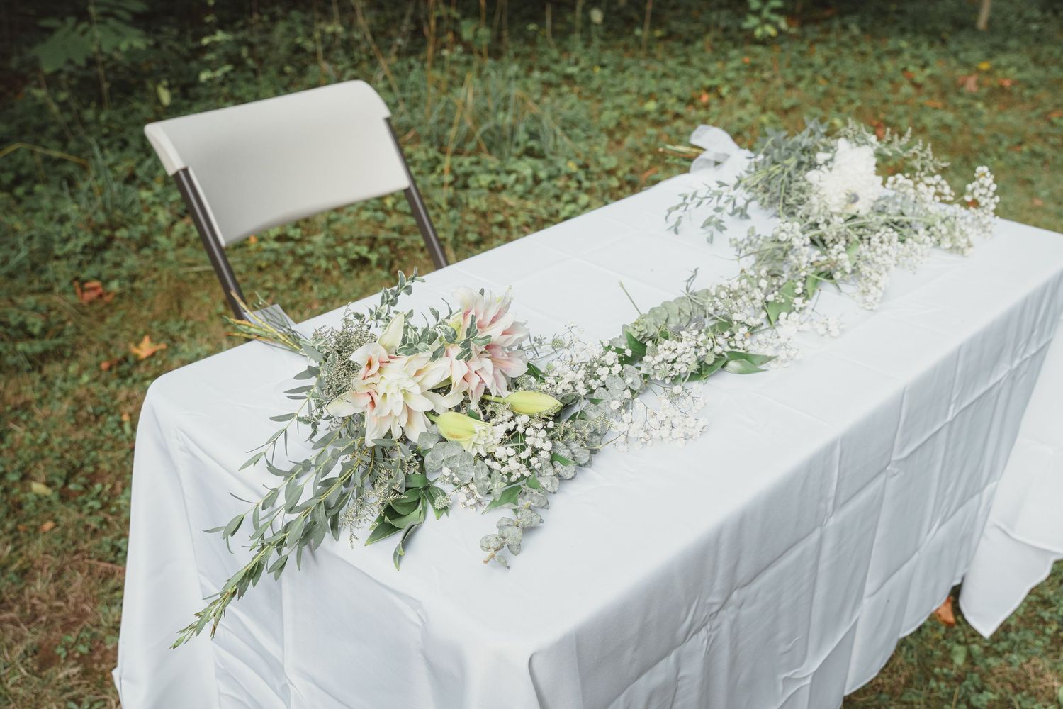 White tablecloth wedding table adorned with elegant floral arrangement of pink flowers and greenery outdoors.