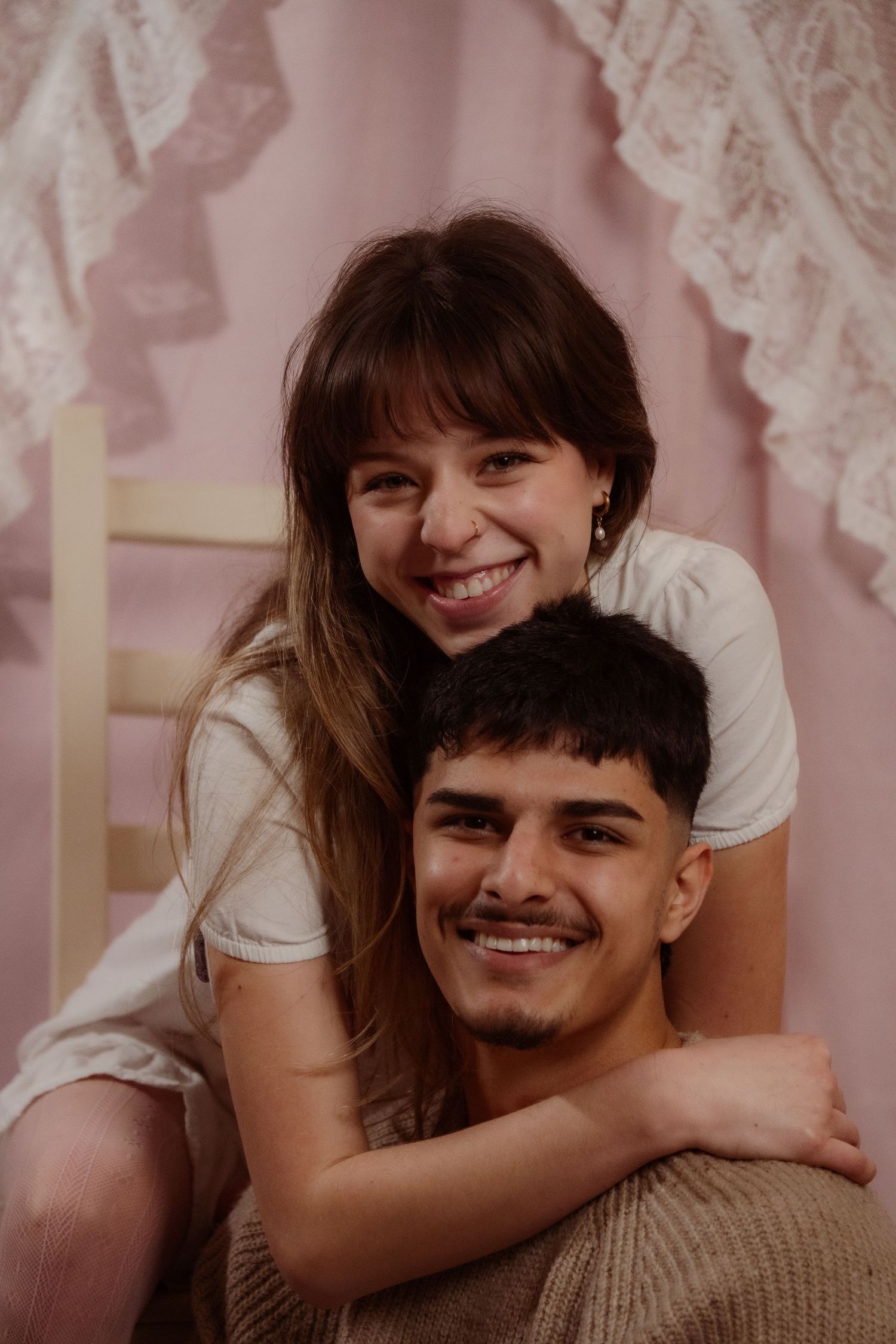 Happy young couple smiling together, woman hugging man from behind against a pink backdrop.