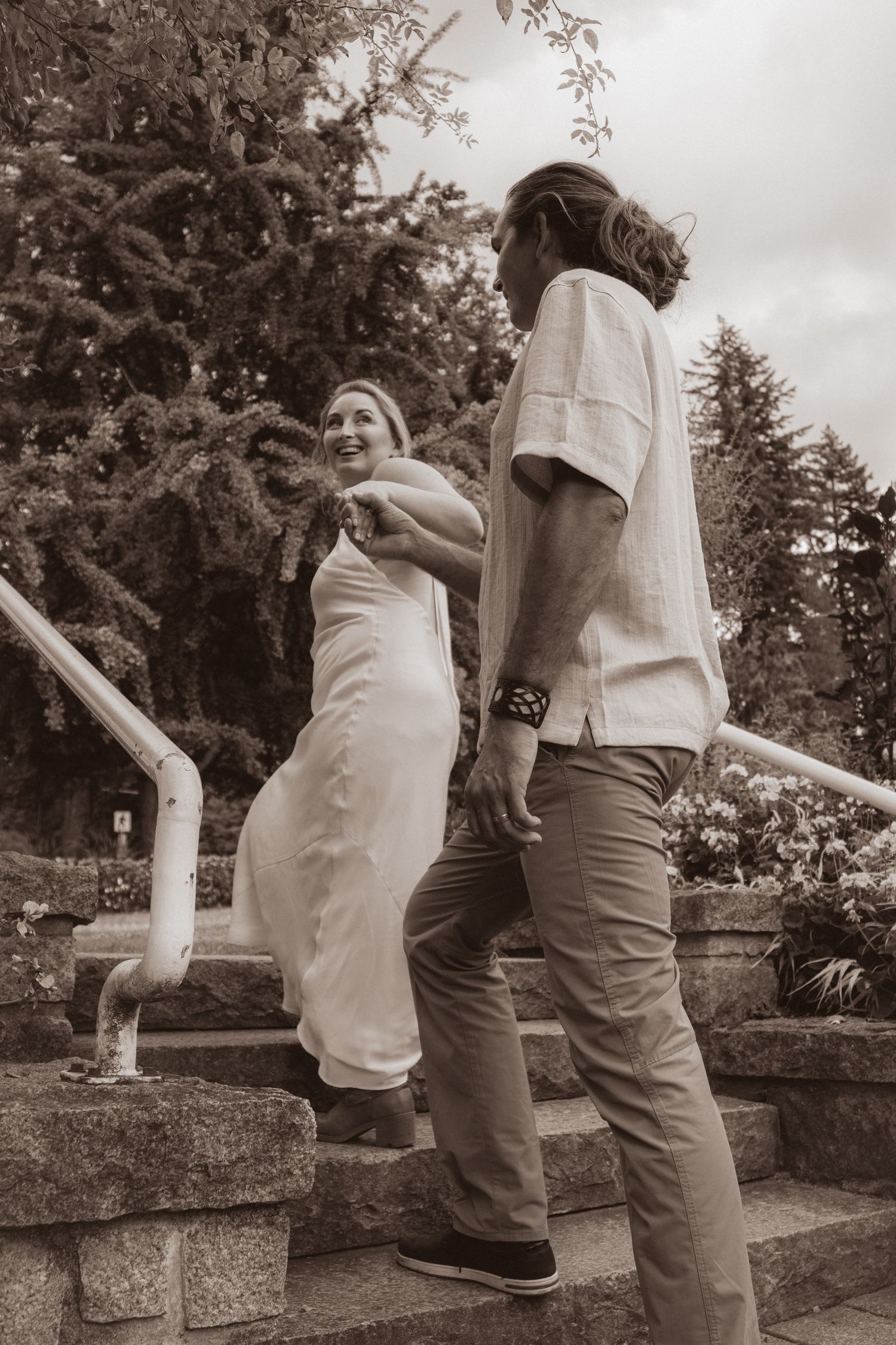 Sepia-toned photo of smiling bride leading groom by the hand up stone garden steps.