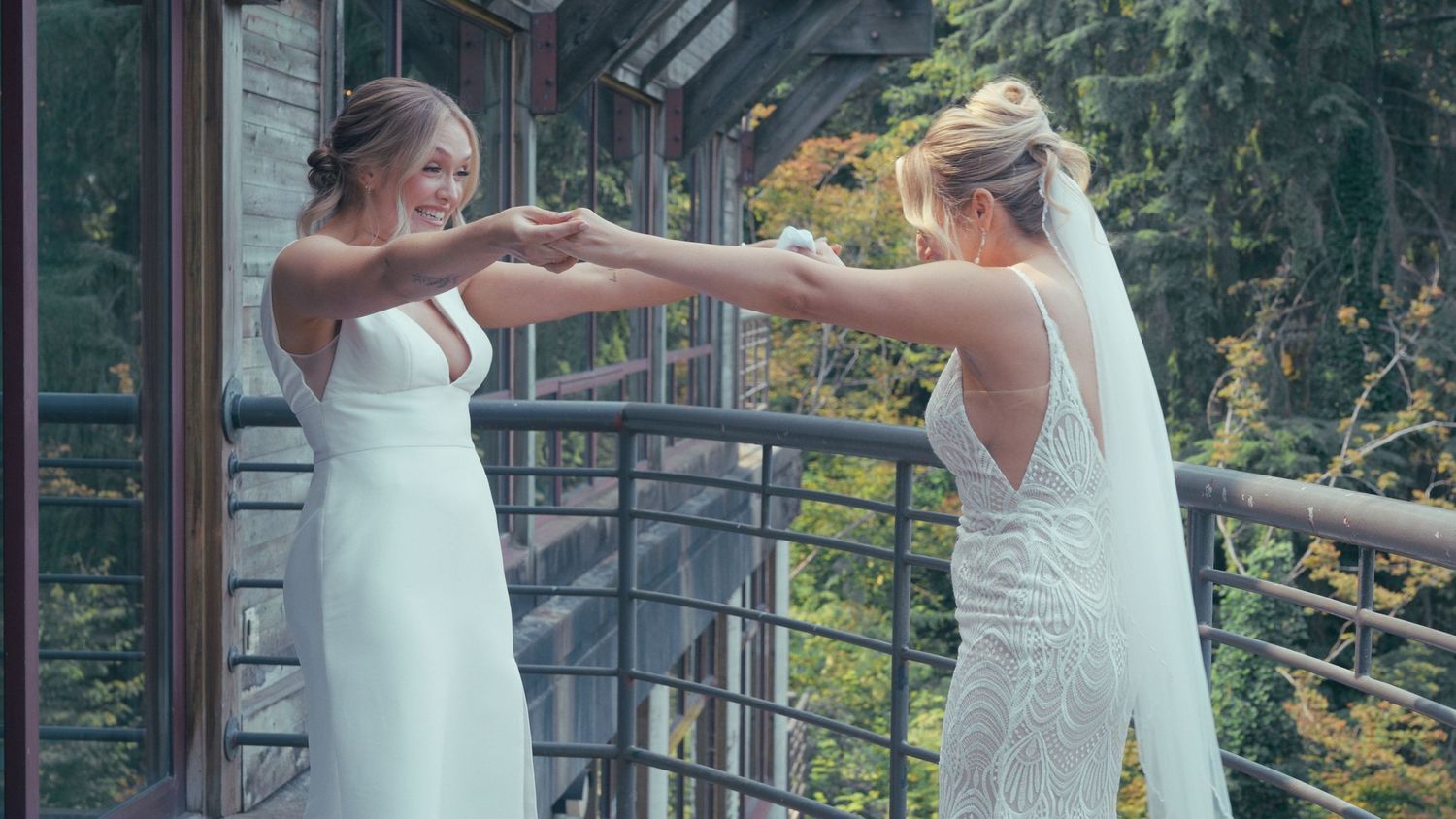 Two brides in white gowns hold hands on balcony surrounded by lush green trees.