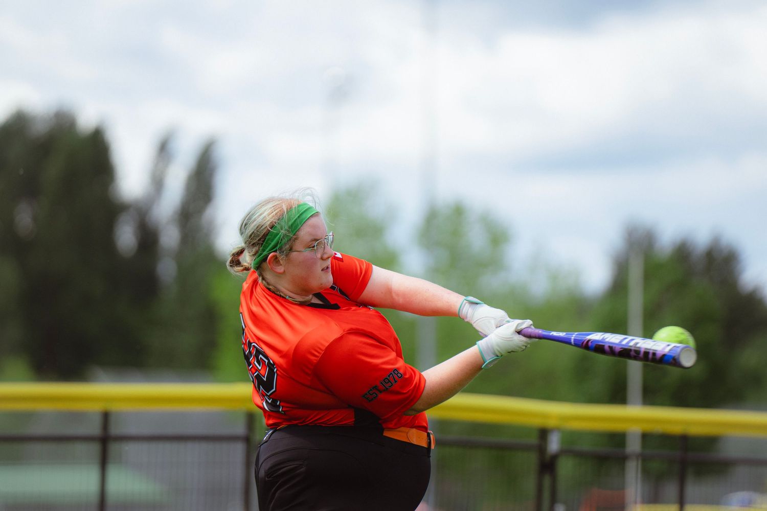 Female softball player in orange jersey swinging bat during outdoor game.