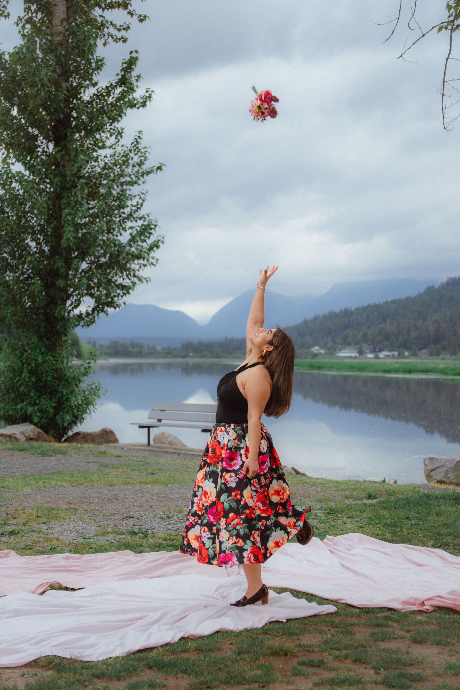 Woman in floral skirt tossing a bouquet into the air by a calm mountain lake.
